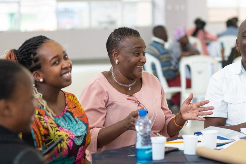 Two women sitting at a round table with others. They are smiling and joyous in appearance.