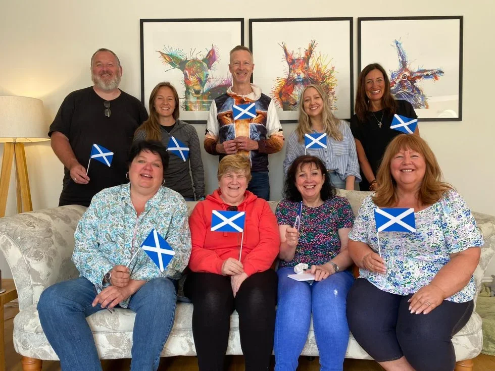4 people on a couch, 5 people standing behind it, all smiling and holding up small Scotland flags in their hands.