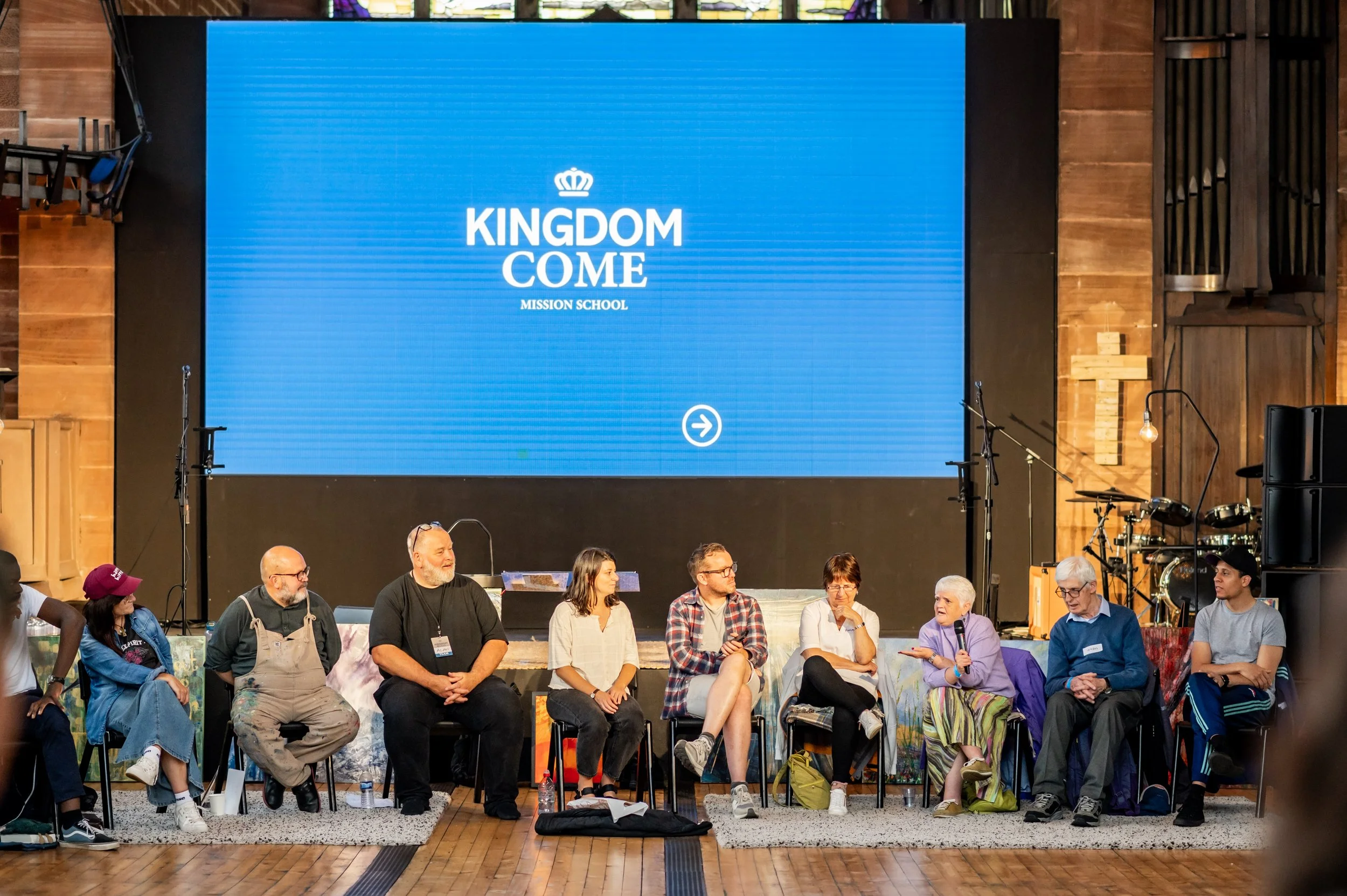 A panel of people sitting on front of a screen saying Kingdom Come, looking towards the one on the panel who is answering into a microphone.