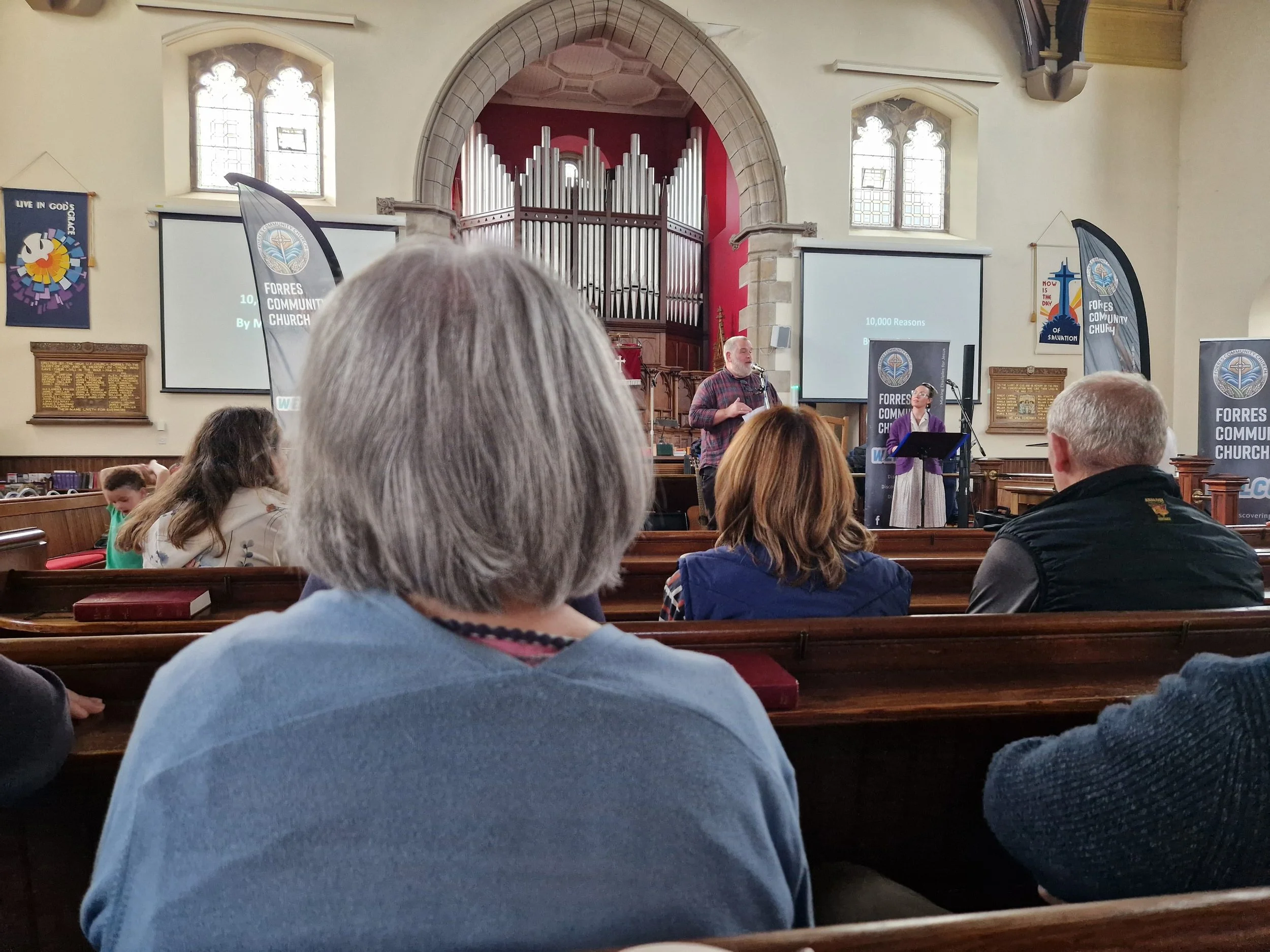 A person sitting in a church pew has taken a photo looking towards the speaker. The backs of peoples heads are seen from the angle of the shot.