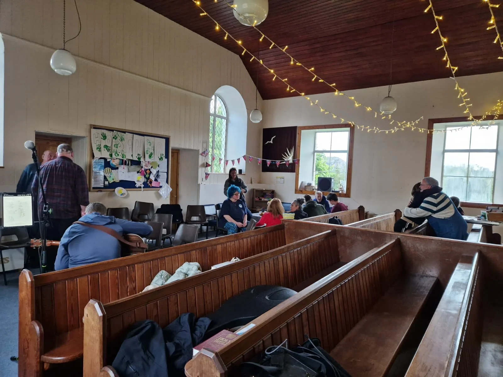 A group of people hanging out in a church, chatting, hugging, someone playing a guitar.
