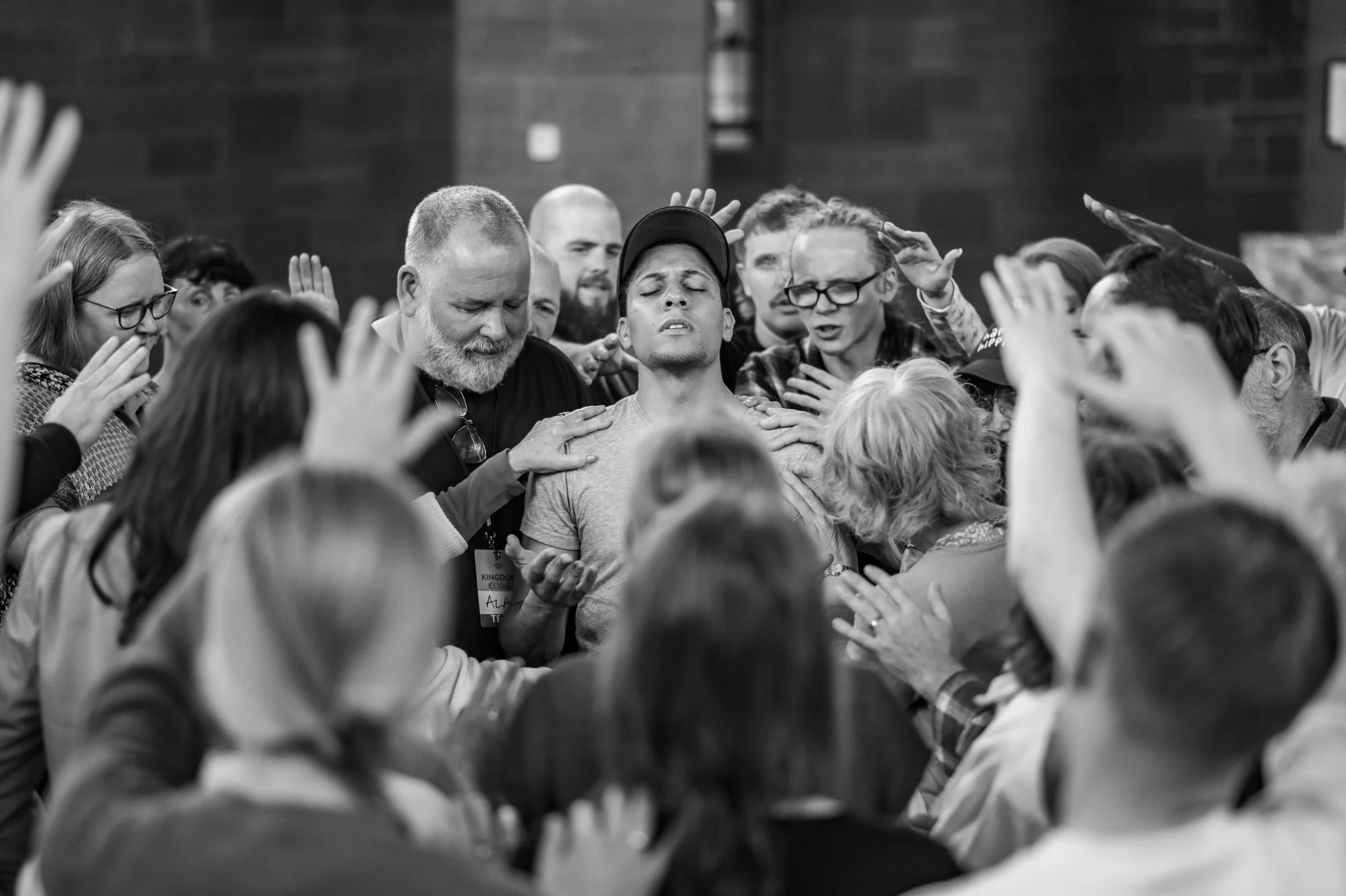 A man in the midst of a large group with his eyes closed and hands lifted, receiving prayer from everyone in the shot.