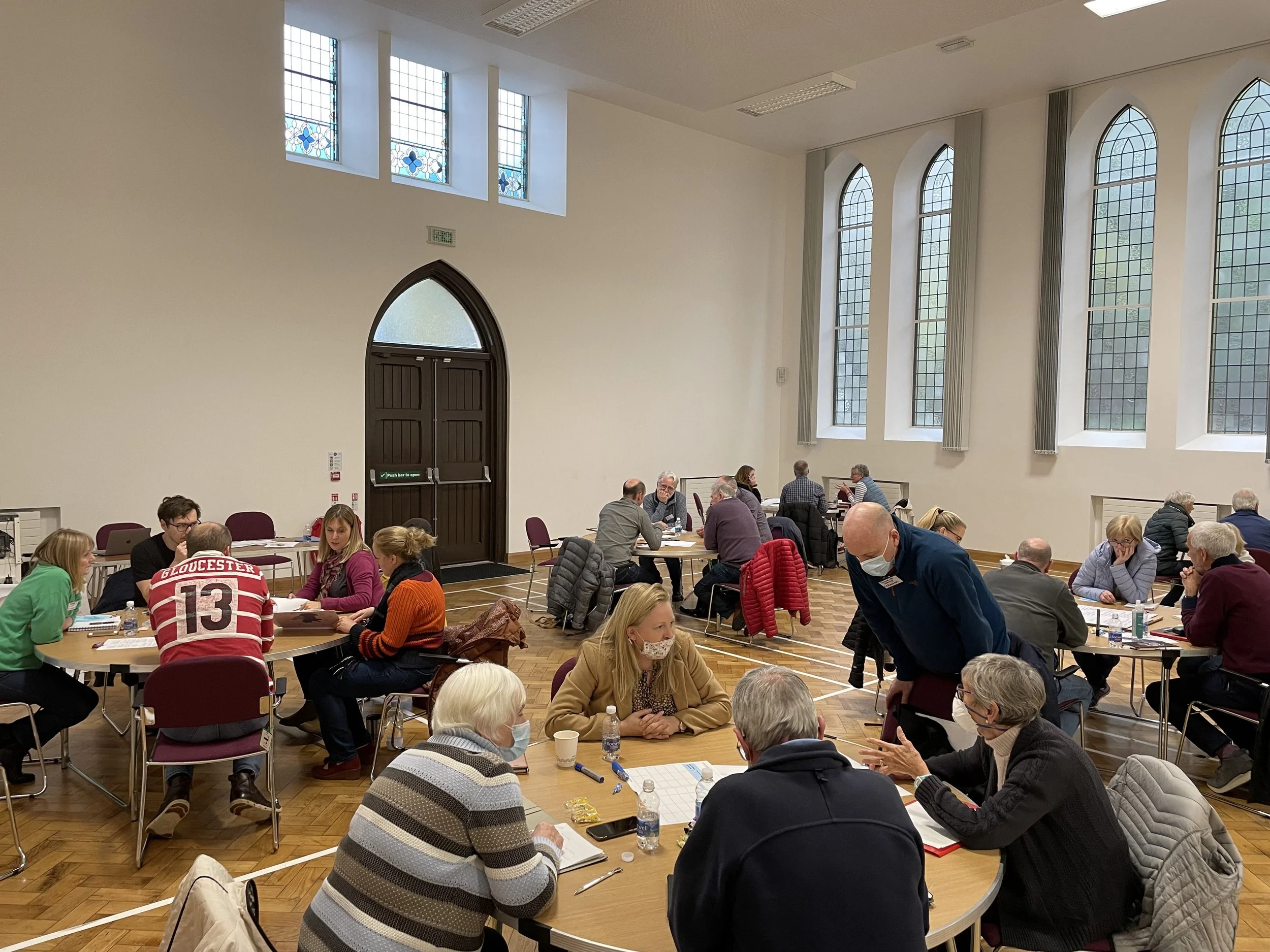 There are round tables spread throughout a large room with groups of people sitting at each one talking within their groups.