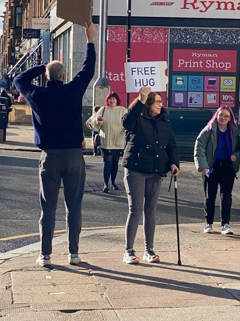Two people holding up signs offering free hugs to folks walking along the street.
