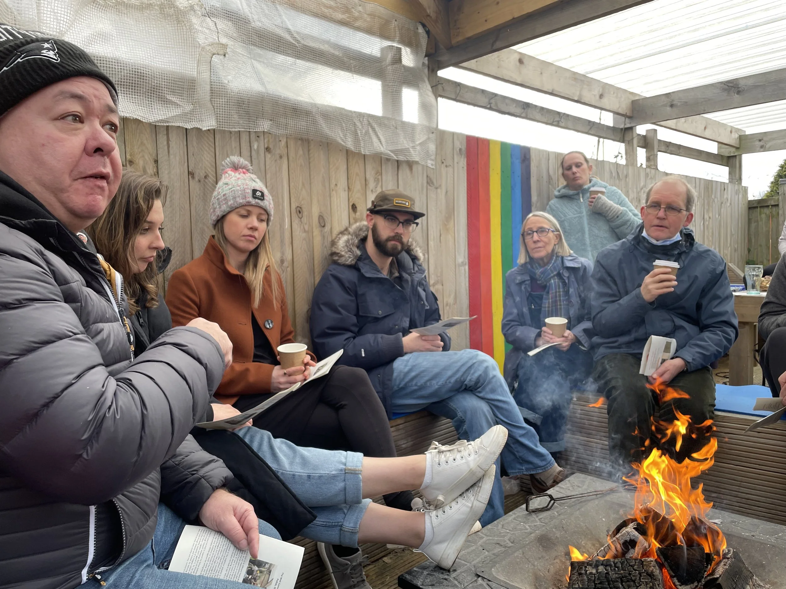 A group of individuals sitting around a fire, holding coffee cups.