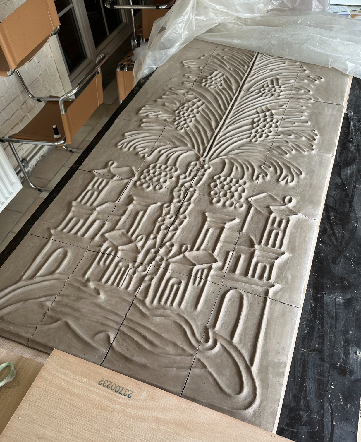 Carrelage en argile avec un motif gravé représentant un arbre avec branches, feuilles, fleurs et des bâtiments en arrière-plan, disposé sur une table.
