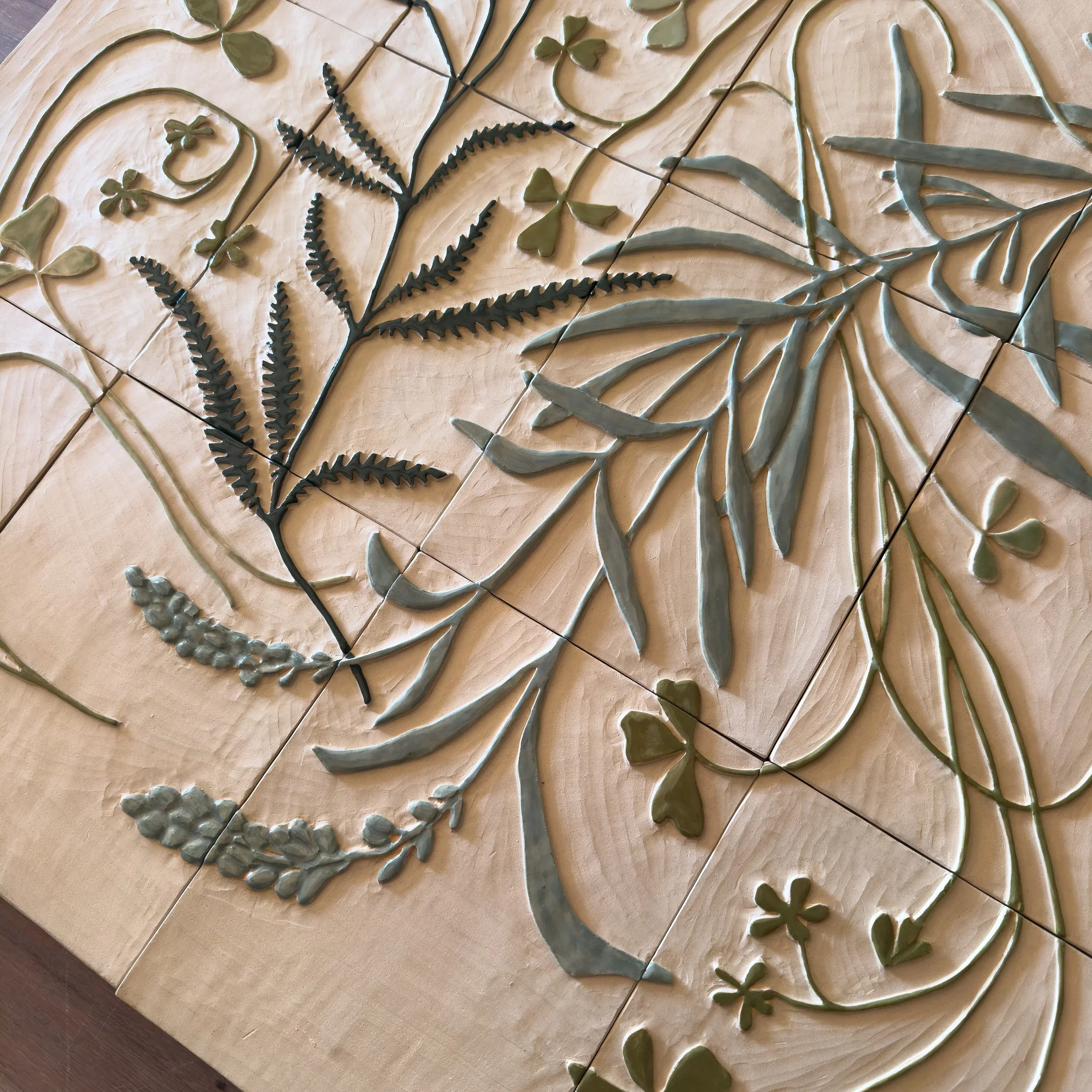 A carved wooden surface featuring a floral design with leaves, stems, and small flower and fern-like details, created with different shades of green and black accents.