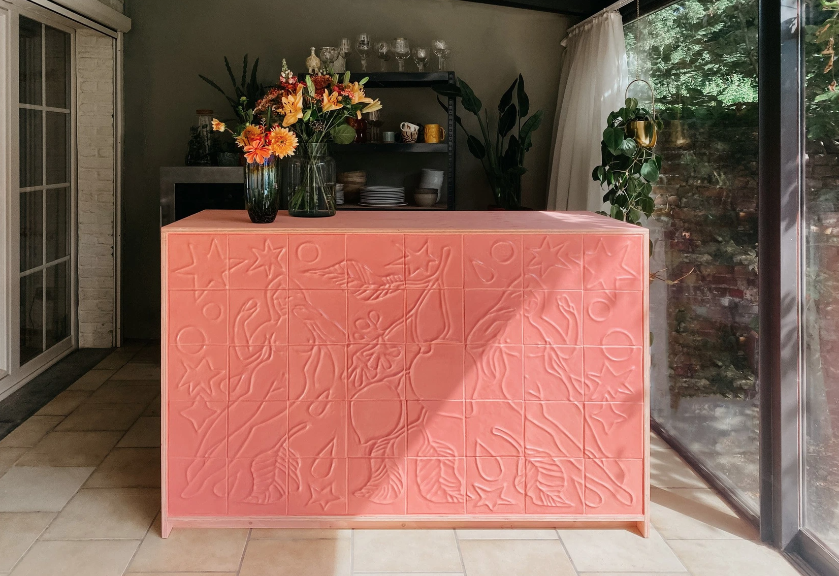 Une table de cuisine rose avec un motif floral en relief, située dans une pièce lumineuse avec des plantes et des étagères en arrière-plan.