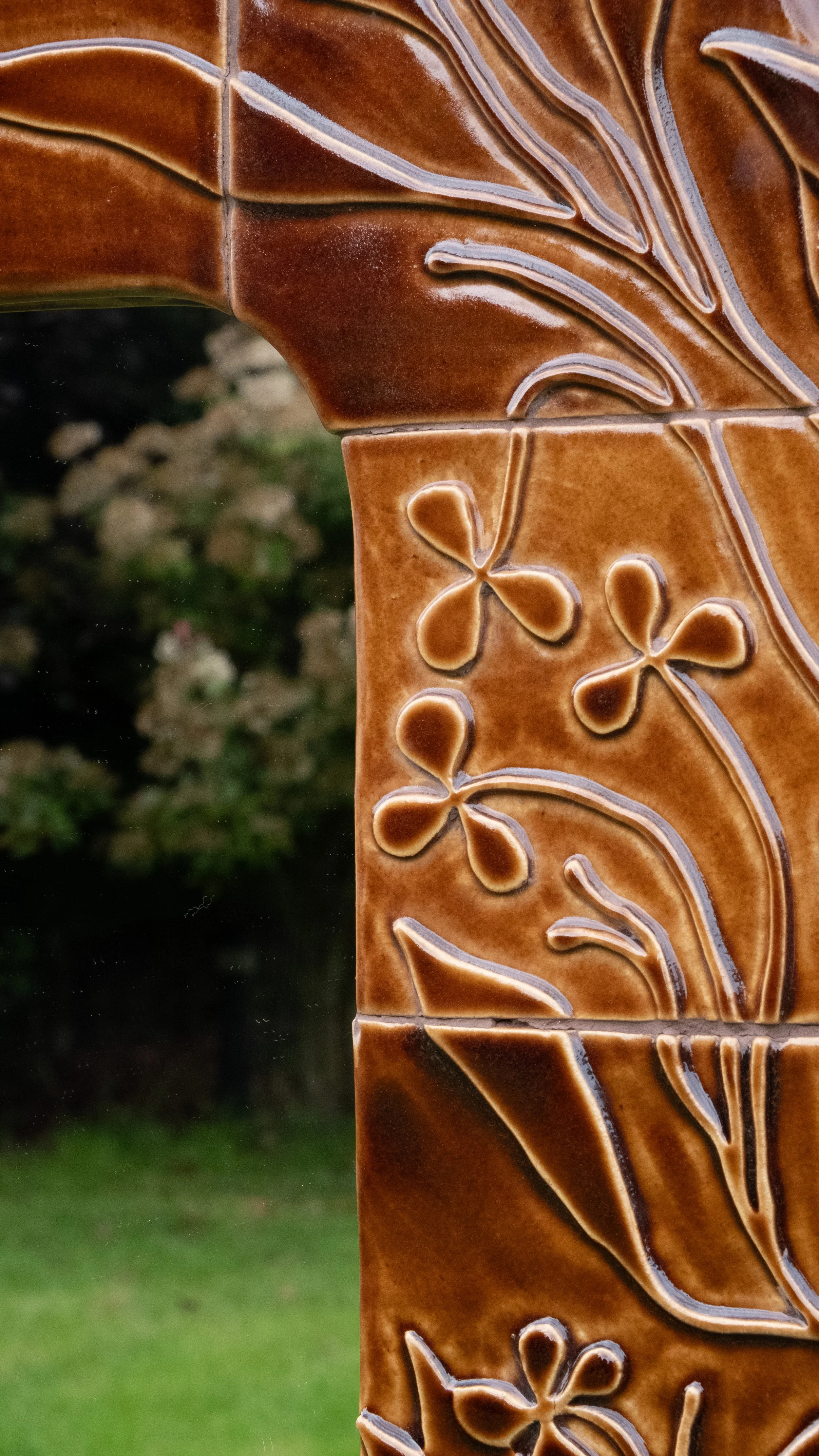 Close-up of a brown ceramic outdoor fountain with carved floral and leaf patterns, with a blurred background of greenery and rocks.
