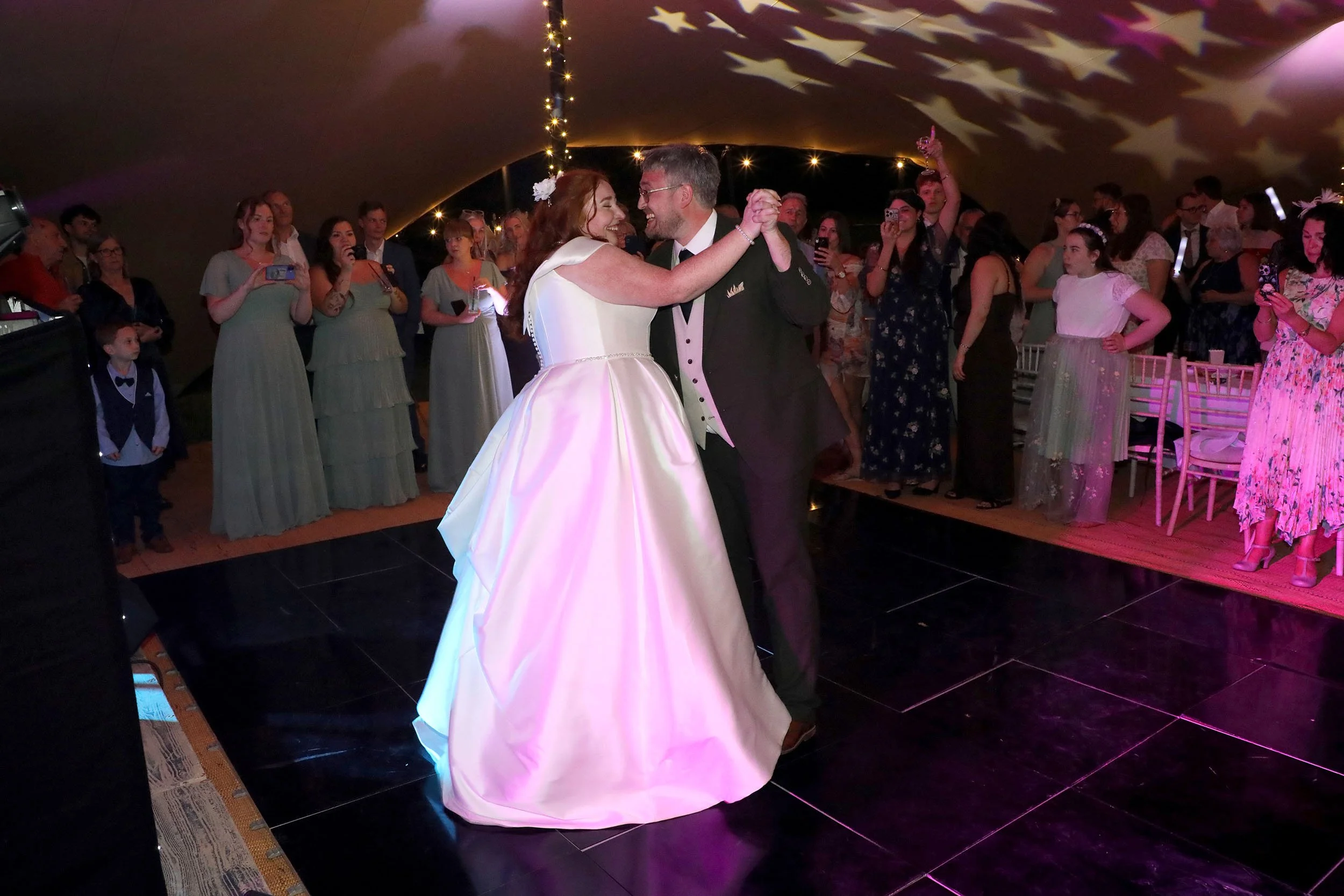 A bride and groom are dancing at their wedding reception indoors, surrounded by guests watching and taking photos. The bride is wearing a white gown with an iridescent sheen, and the groom is in a dark suit with a white shirt. The ceiling has a star-