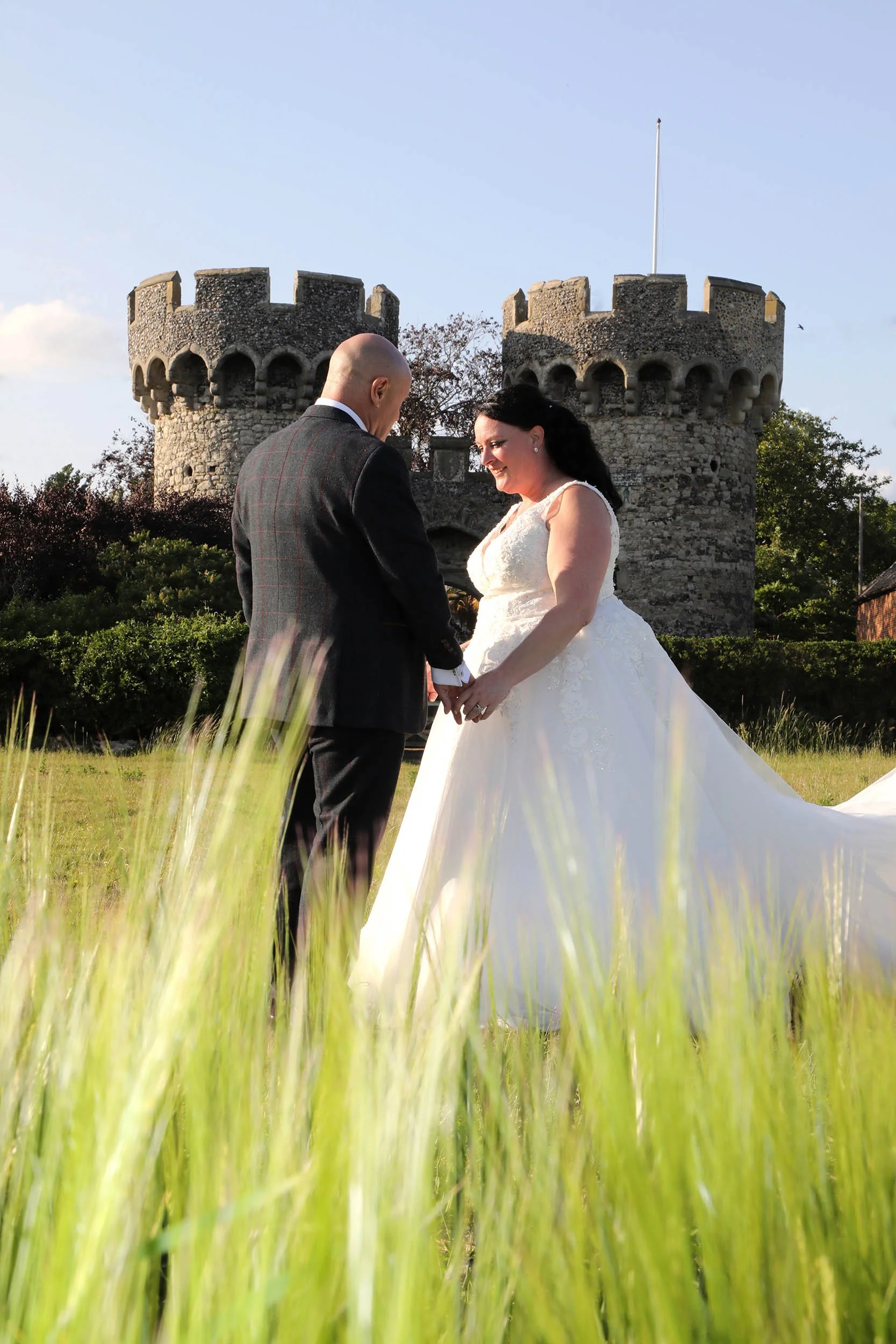 A bride and groom holding hands and smiling at each other outdoors in front of a historic stone castle.