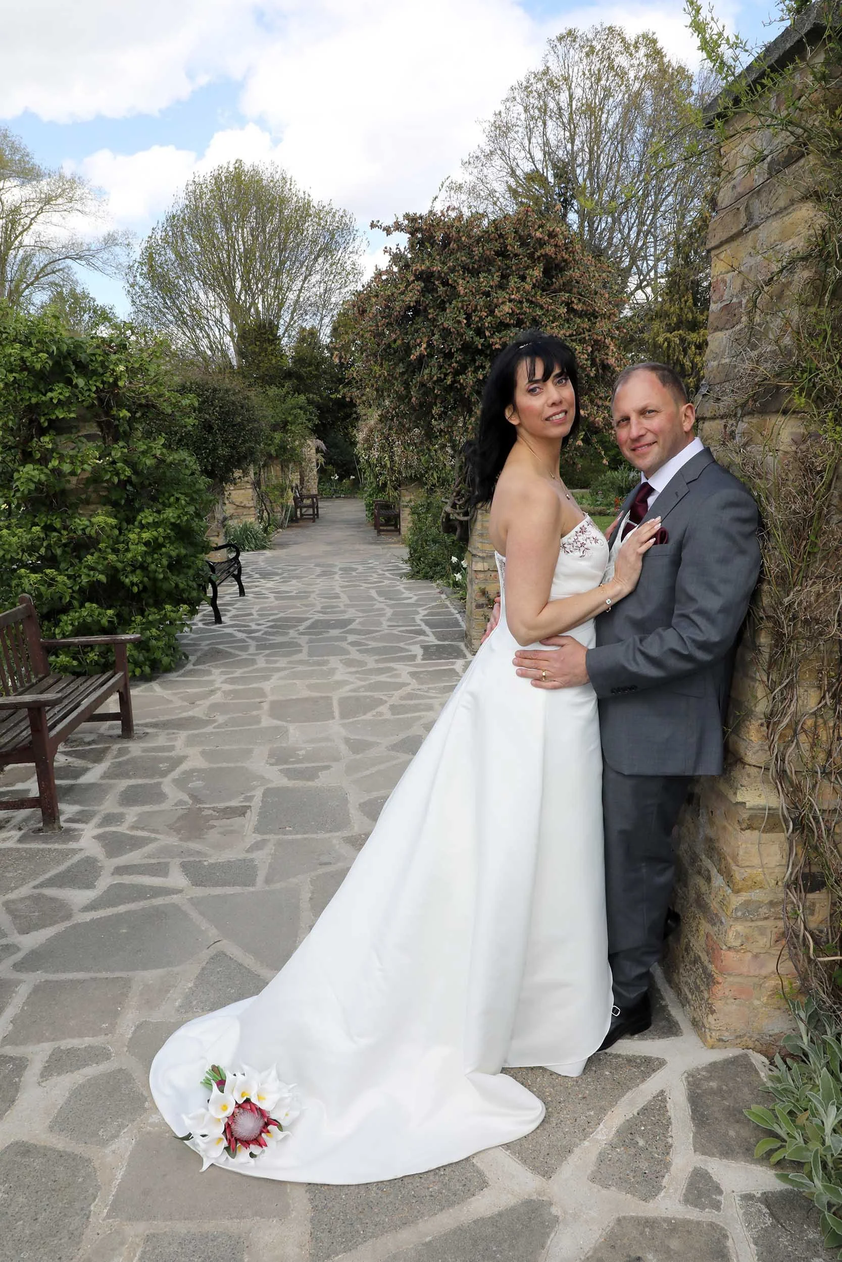 A bride and groom standing close together outdoors on a stone pathway surrounded by trees and bushes, with a cloudy sky overhead. The bride is wearing a white wedding gown with a long train and floral bouquet at the bottom, and the groom is in a gray