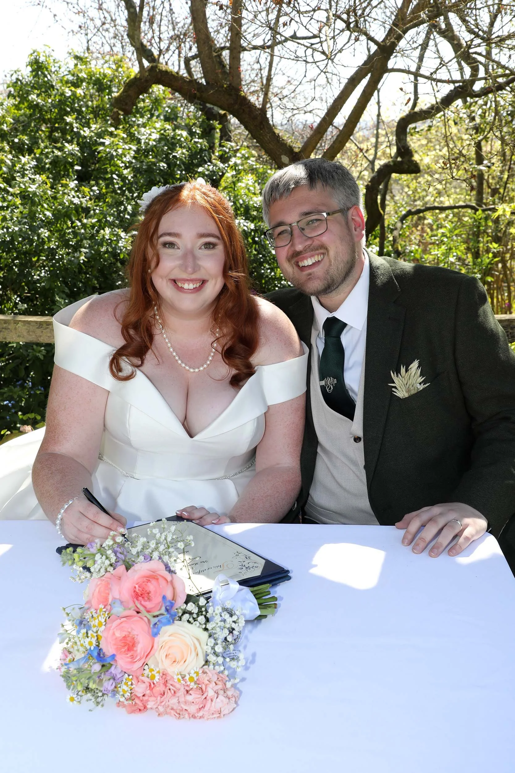 A bride and groom smile at a wedding ceremony outdoors. The bride has red hair, is wearing a white off-shoulder wedding dress, pearl necklace, and floral headpiece. The groom has glasses, short gray hair, and is wearing a black suit, white shirt, and