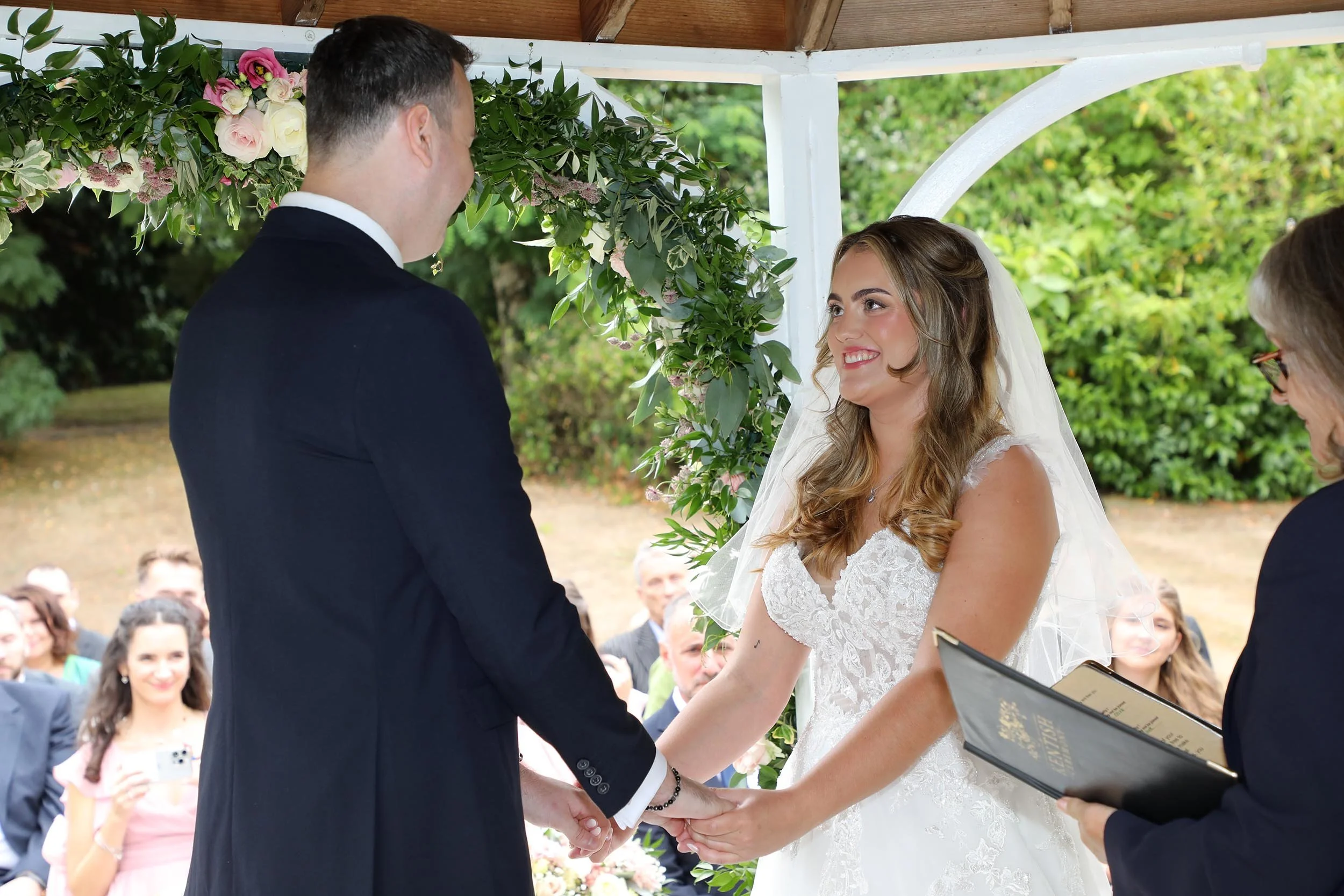 A bride and groom holding hands during their wedding ceremony under a decorated arch, with guests seated in the background.