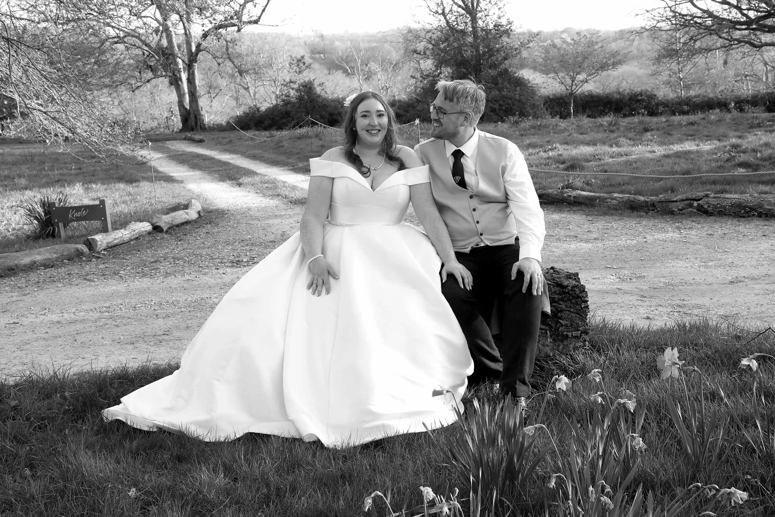 A black and white photo of a bride and groom sitting outdoors on a dirt path with trees and hills in the background. The bride wears a white wedding gown and the groom wears a white shirt with a tie. They are smiling and looking at each other.