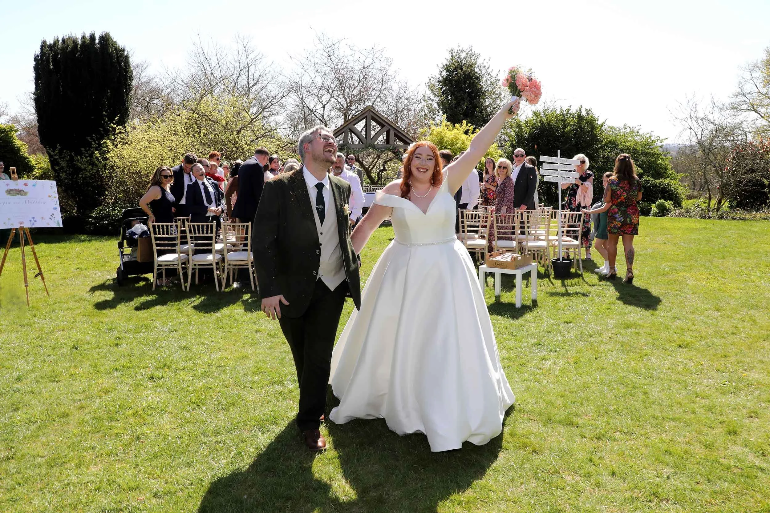 A bride and groom are celebrating outdoors at a wedding, with the bride holding a bouquet of pink flowers and raising her arm. Guests are in the background, some smiling and others looking on. The setting is a sunny garden with chairs arranged for th