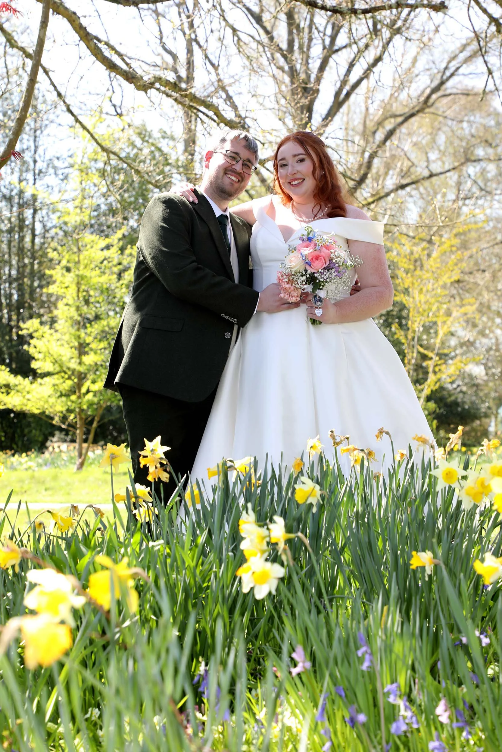 A bride and groom smiling outdoors during daytime, standing behind yellow and purple flowers with green grass, trees in spring foliage, and sunlight creating a bright background.