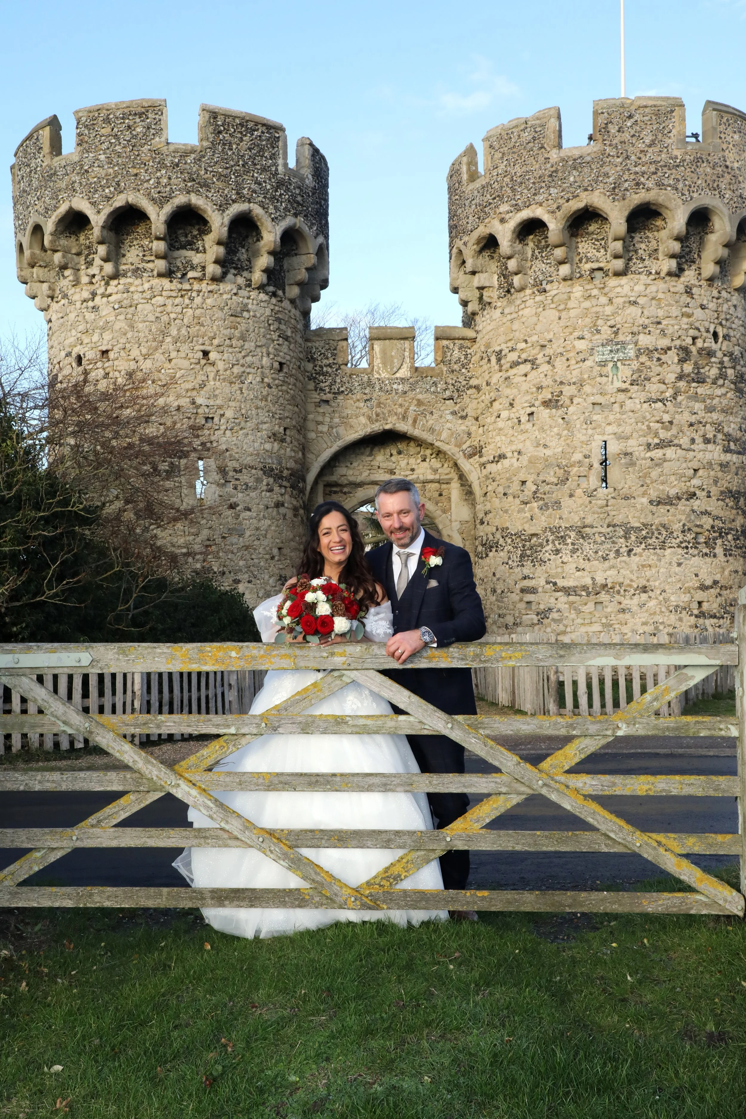 A bride and groom smiling and posing behind a wooden fence in front of Cooling Castle towers.