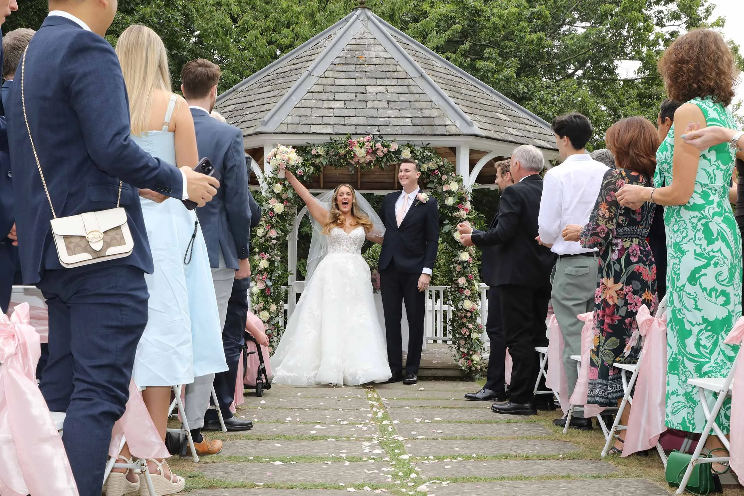 A wedding ceremony outdoors with a bride and groom standing under a floral arch, surrounded by seated guests on either side, on a cloudy day with green trees in the background.