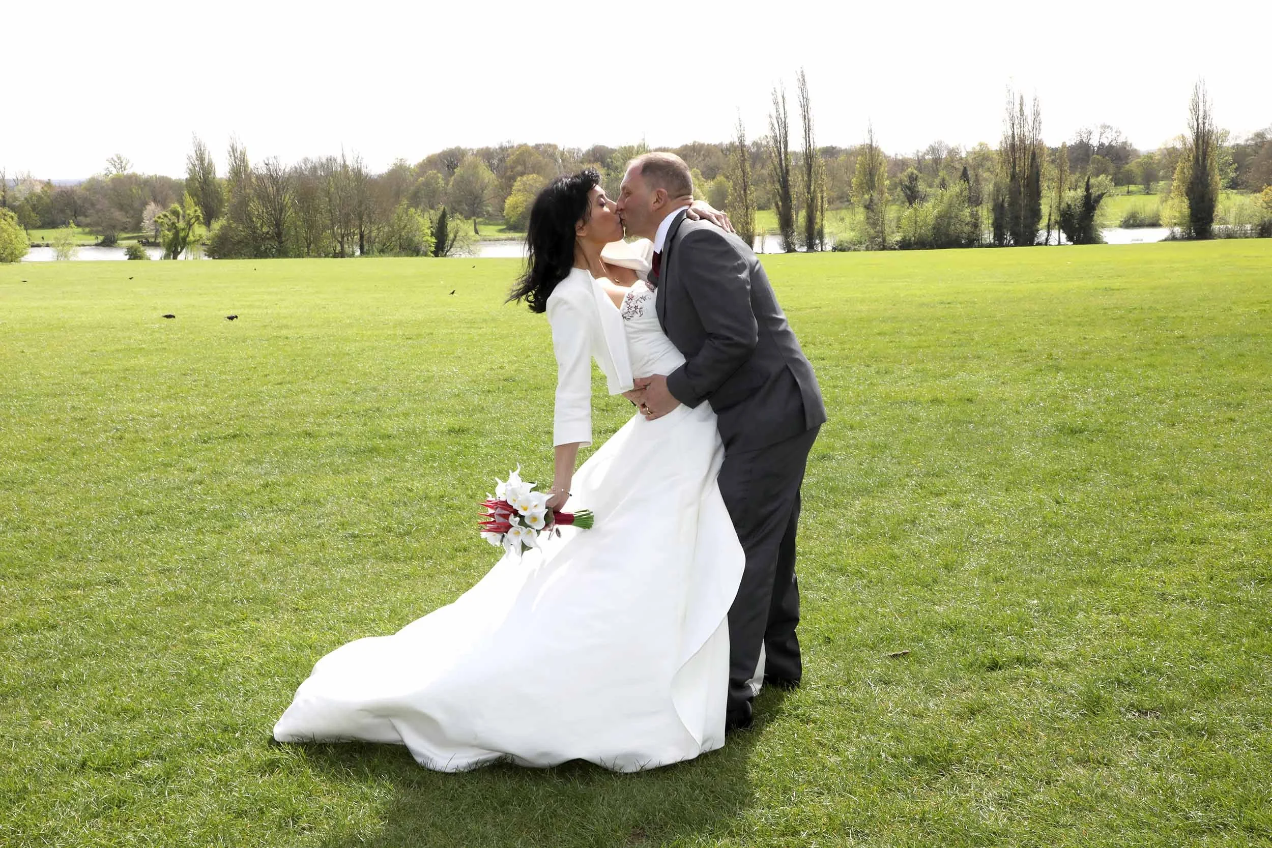 A man and woman dressed in wedding attire sharing a kiss in an open grassy field with trees and a pond in the background.