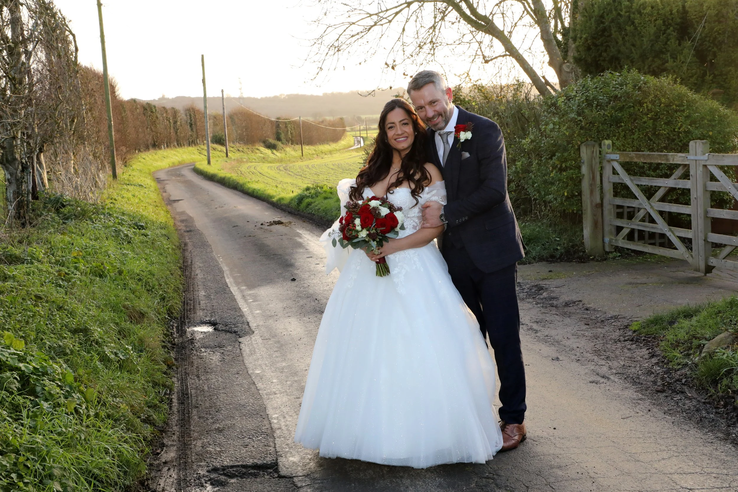 A newlywed couple stands on a rural road during sunset, smiling and holding a bouquet of red and white flowers. The bride wears a white wedding gown, and the groom wears a dark suit. The background features a winding road, trees, and a wooden gate.