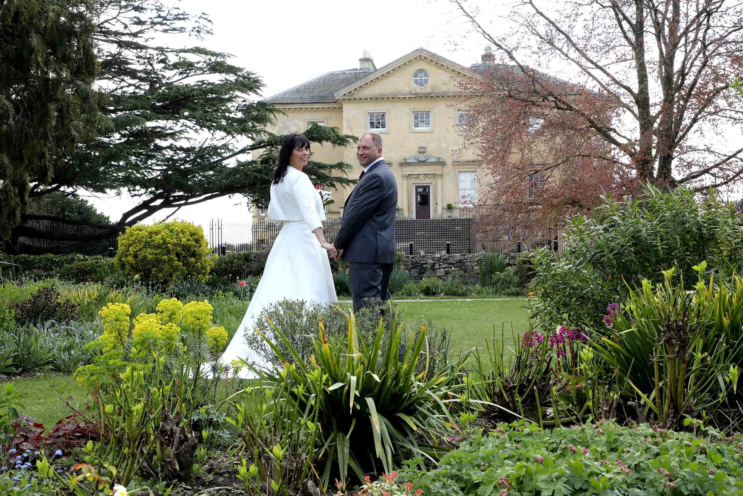 A newlywed couple holding hands and smiling in a garden with colorful flowers and green shrubs, with a large yellow house and trees in the background.