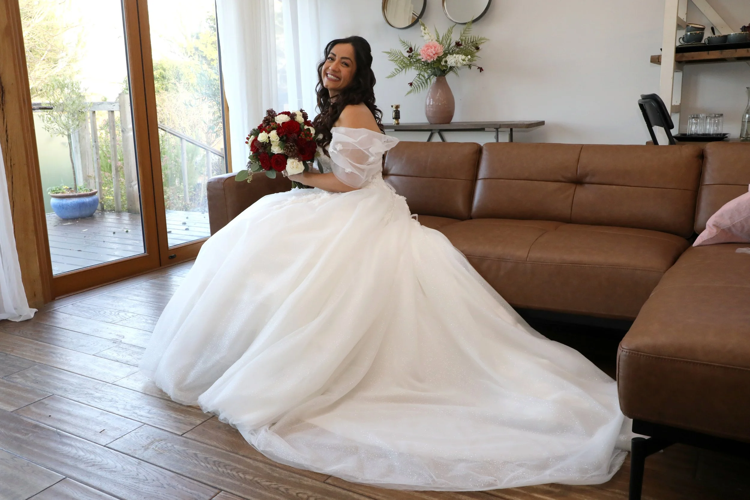 A smiling woman in a wedding dress holding a bouquet of red and white flowers sits on a brown leather couch in a bright room with large glass doors, a flower arrangement on a table, and a bookshelf with dishes and glasses.