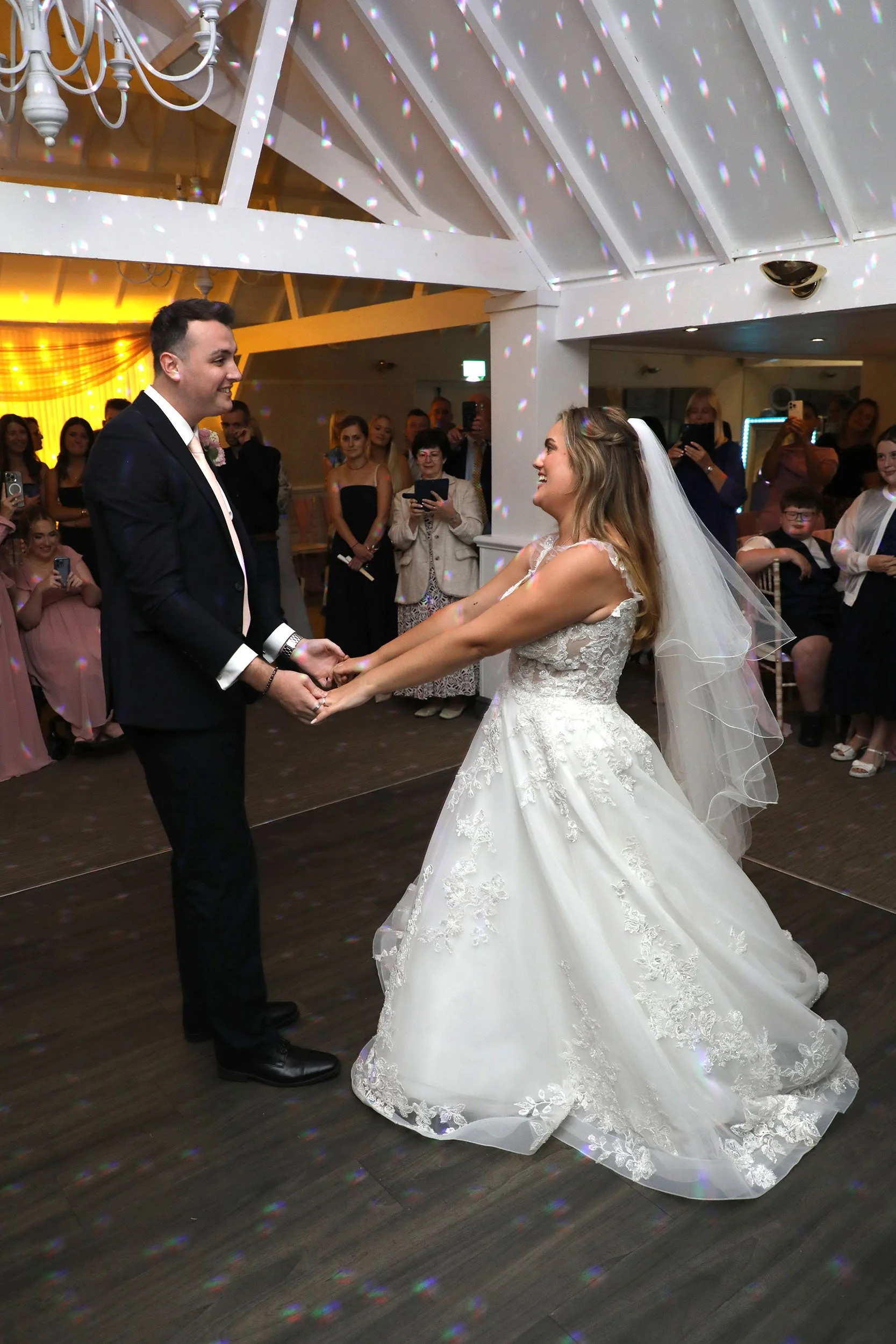 A bride and groom are holding hands during their wedding ceremony, surrounded by guests. The bride is in a white wedding dress with lace details and a veil, and the groom is wearing a black suit. The scene is decorated with colorful lights reflecting