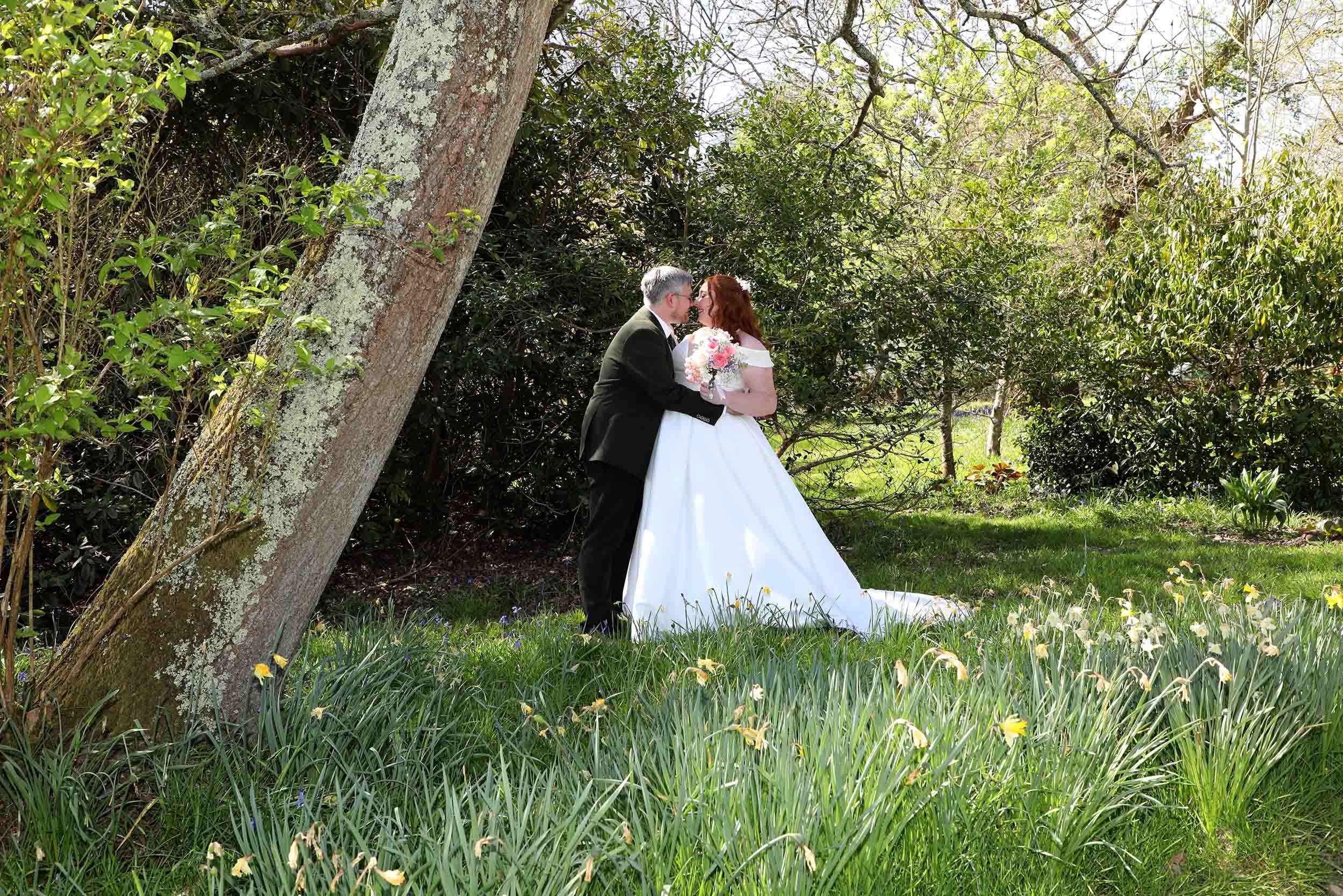 A bride and groom sharing a kiss outdoors beneath trees in a grassy, floral setting.