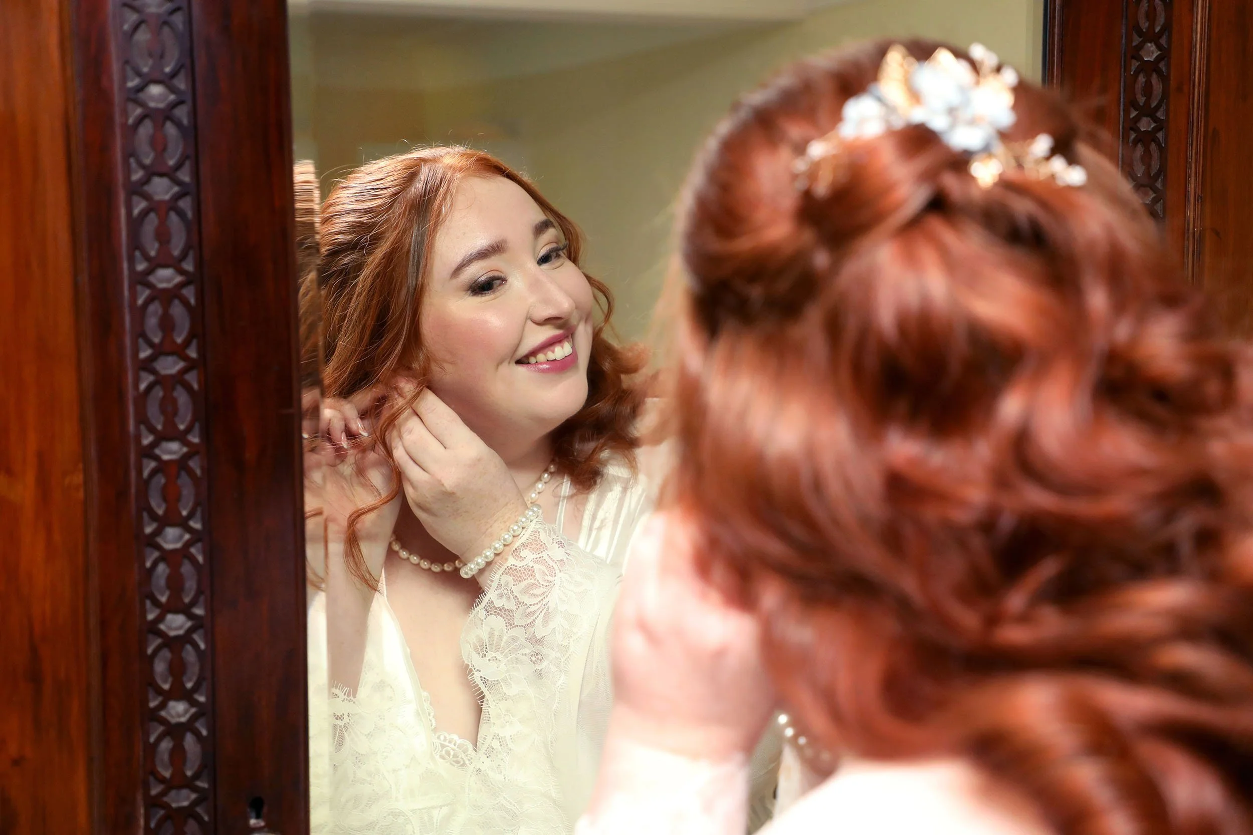 A woman with red hair and a pearl necklace is looking at herself in a mirror, smiling, while adjusting her earring. The reflection shows her lace dress and elegant makeup, and her hair is decorated with white flowers.
