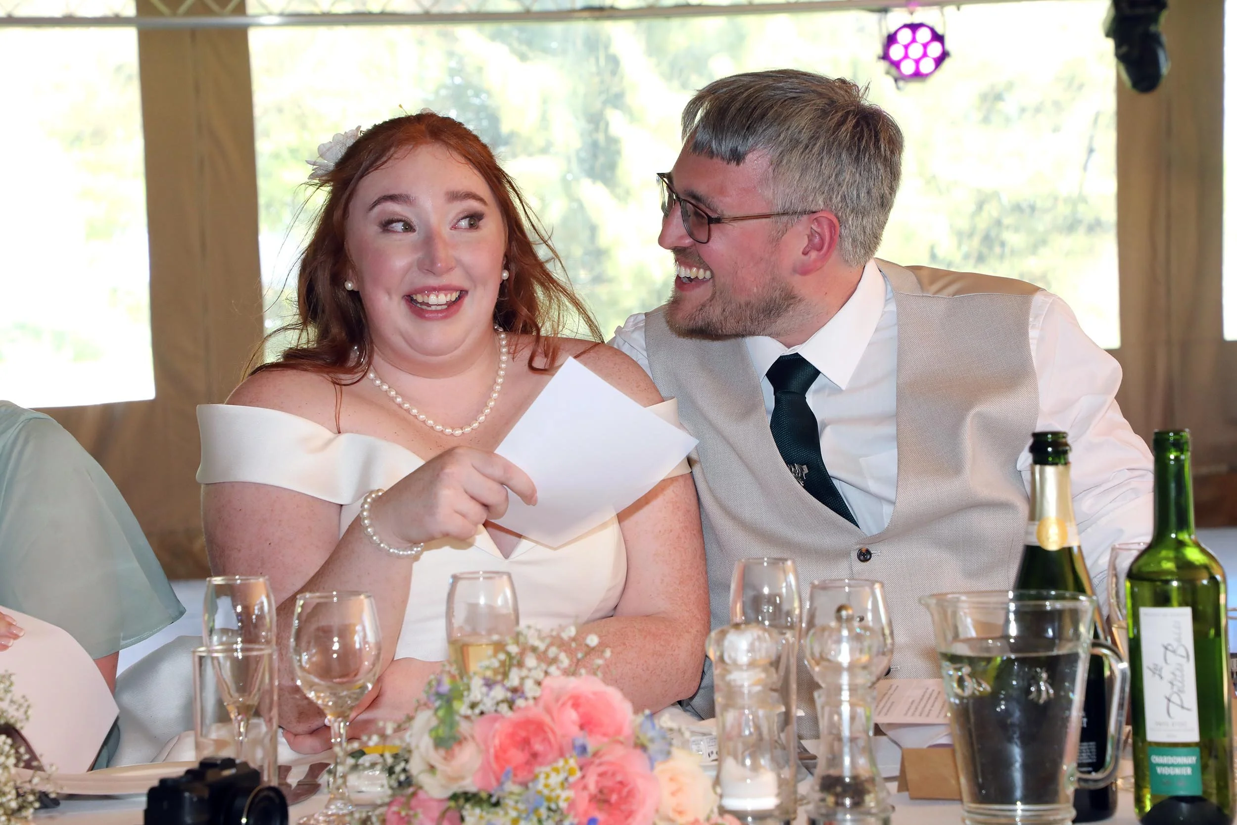 A woman with red hair and a man with glasses are sitting at a table, smiling and looking at each other during a celebration or wedding reception. The woman is holding a piece of paper, and there are glasses, bottles, and floral decorations on the tab