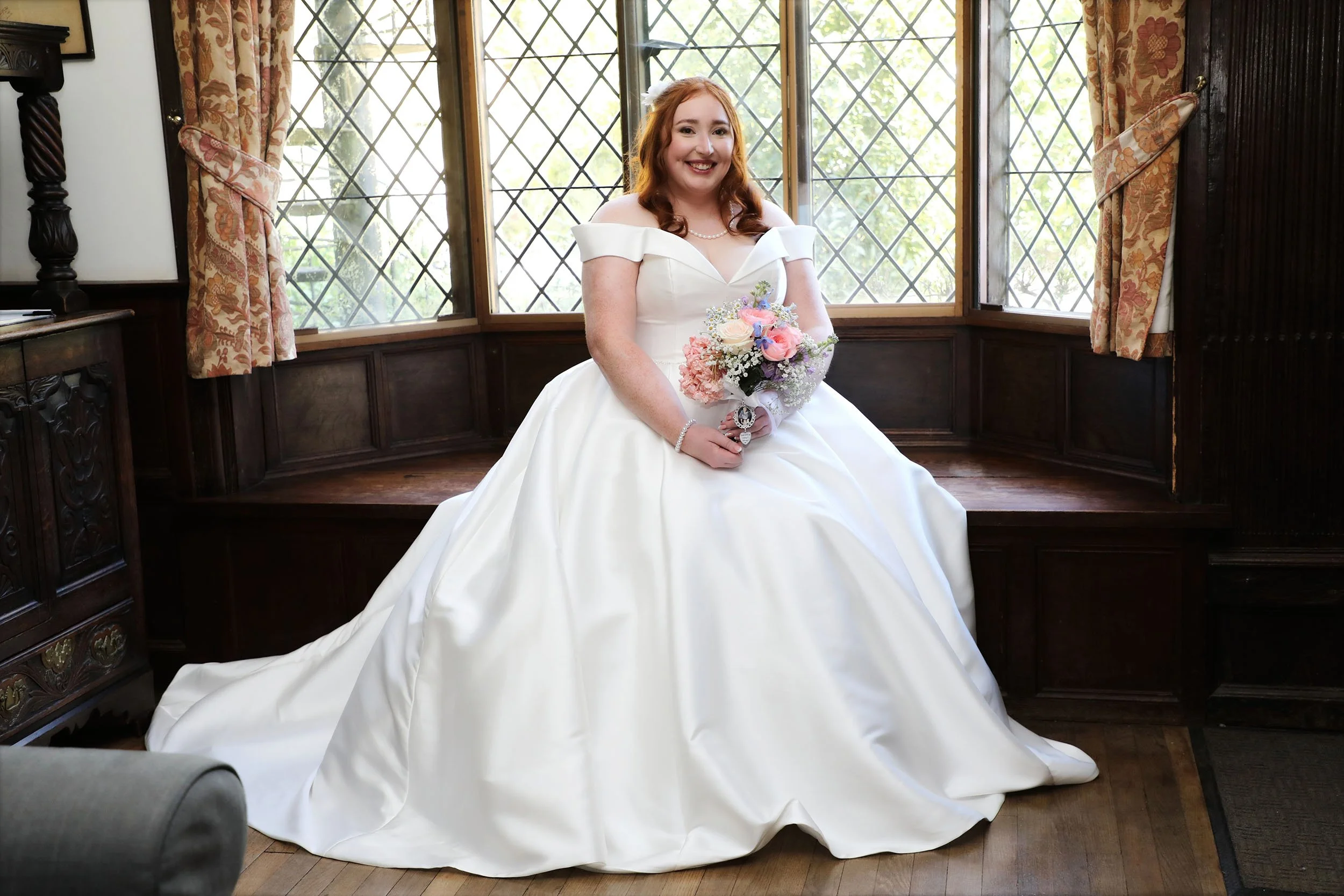 A young woman in a white wedding dress sitting on a wooden window seat, holding a bouquet of pink, purple, and white flowers, smiling inside a room with large lattice windows and floral curtains.