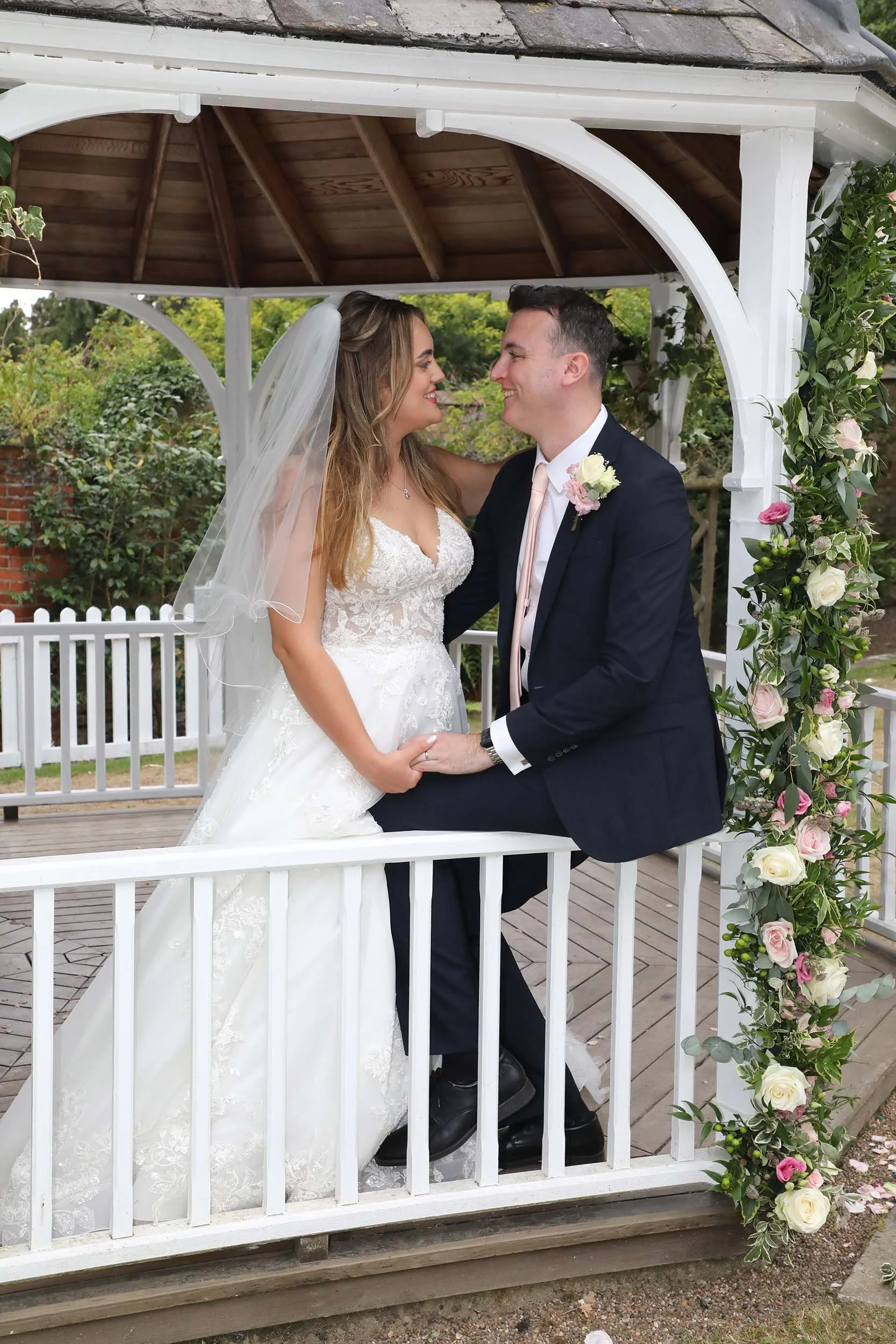 A bride and groom holding hands, smiling at each other, standing beneath a white wooden gazebo adorned with pink and white roses and greenery during their wedding ceremony outdoors.