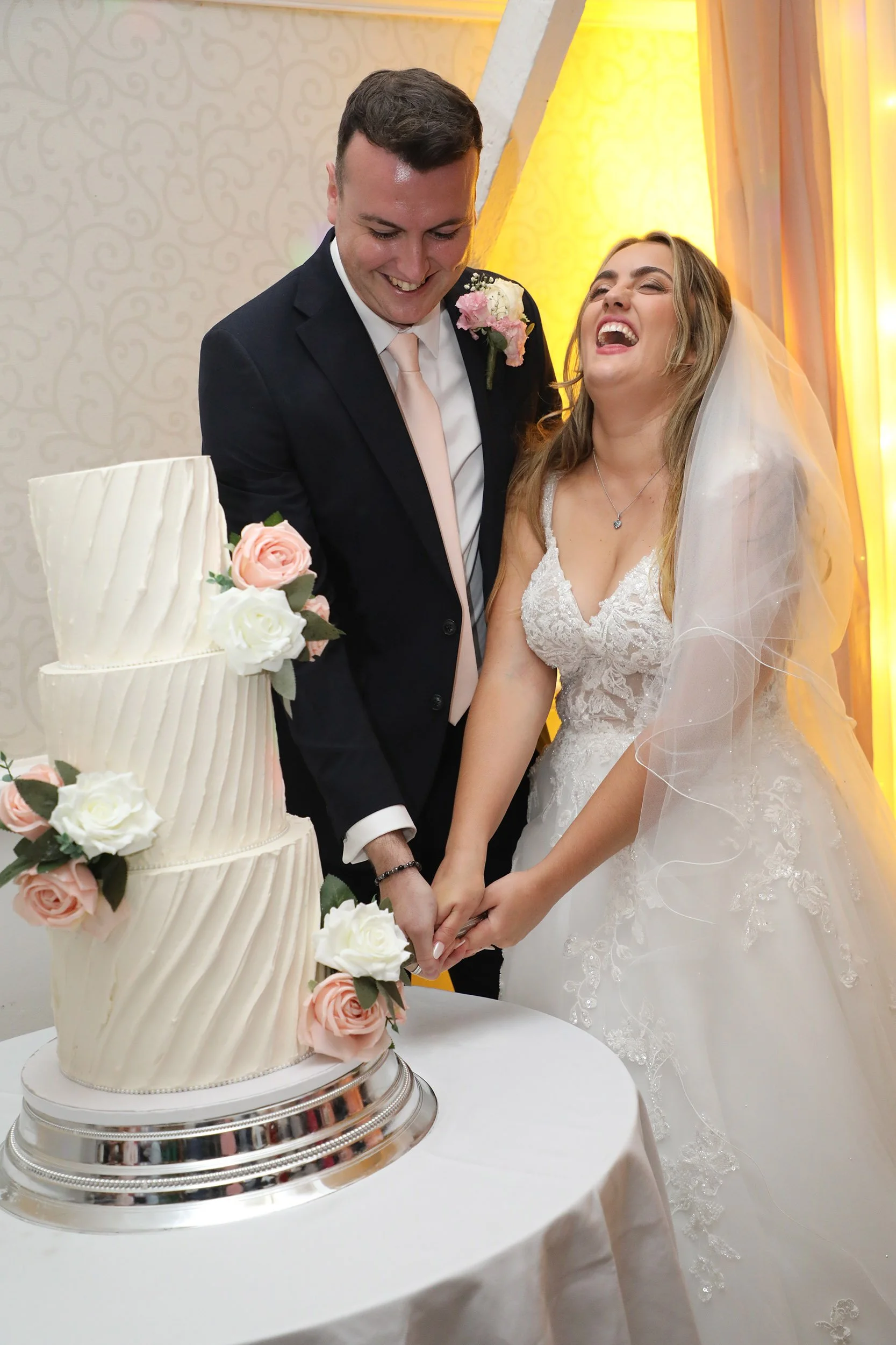 A newlywed bride and groom smiling and laughing while cutting a wedding cake together. The wedding cake is decorated with pink and white flowers.
