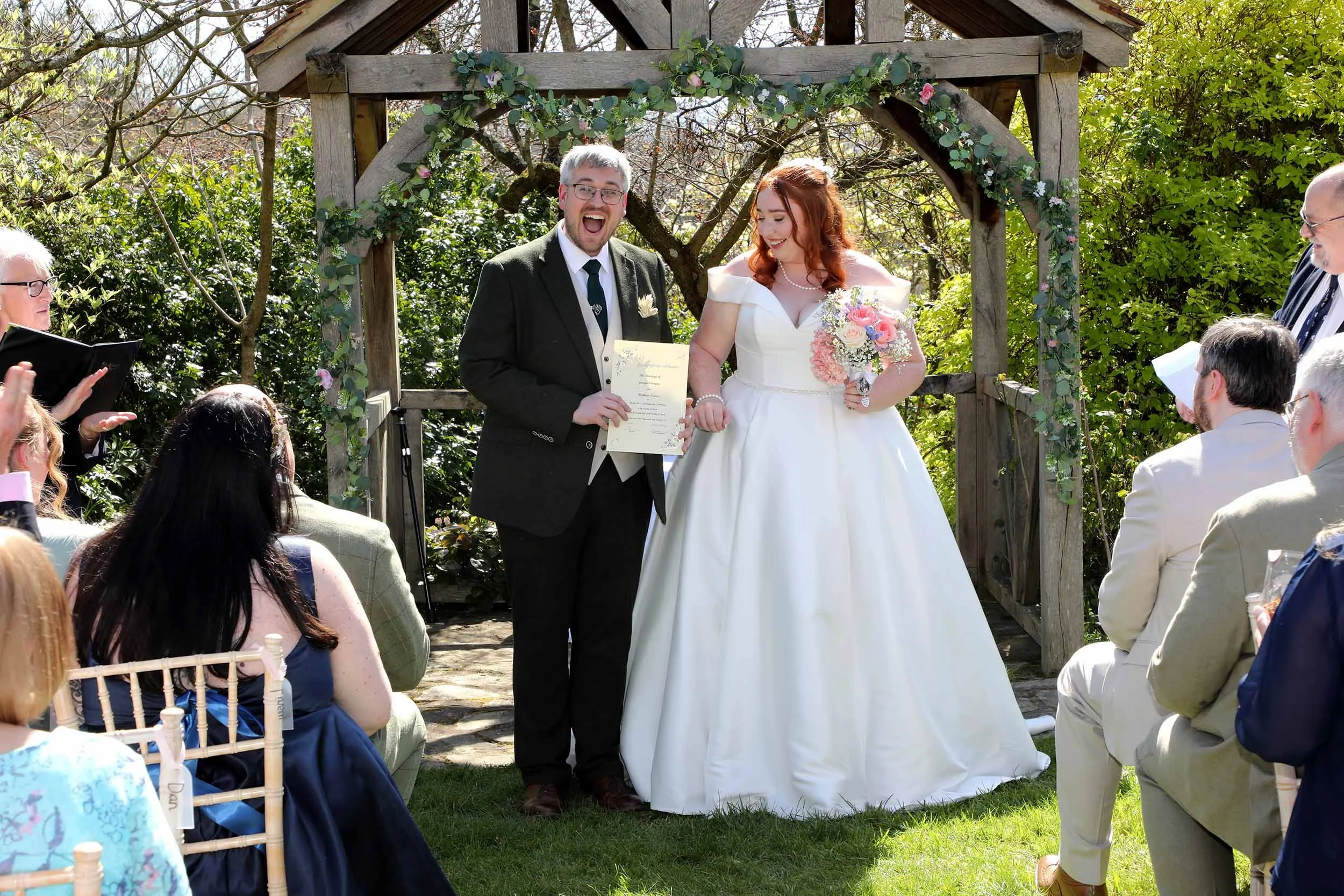 A wedding ceremony outdoors with a bride and groom holding hands under a wooden arch decorated with greenery and pink flowers, surrounded by seated guests in bright sunlight.