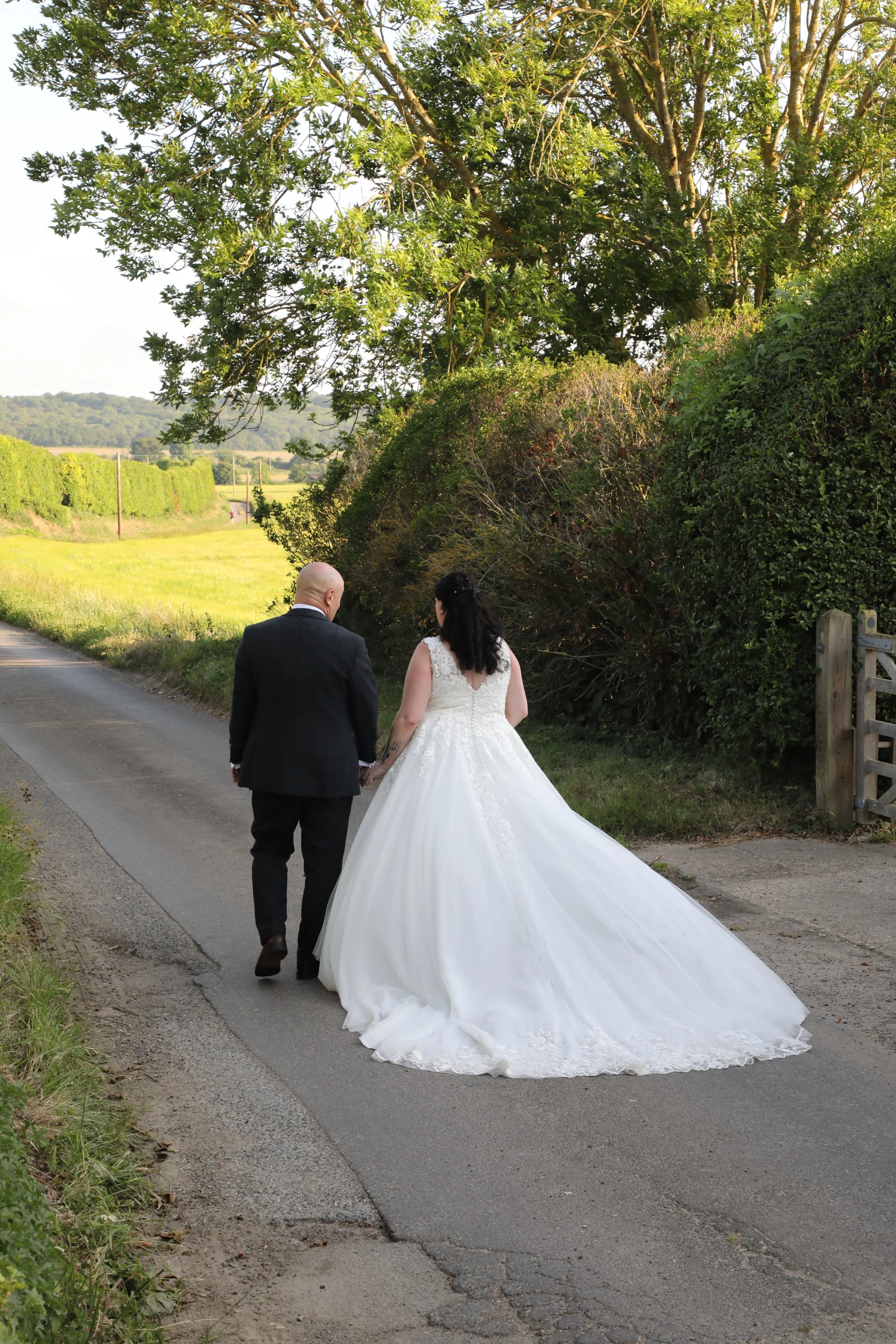 A bride and groom walking hand in hand on a rural country road, surrounded by greenery with a large tree overhead and a scenic landscape in the background.
