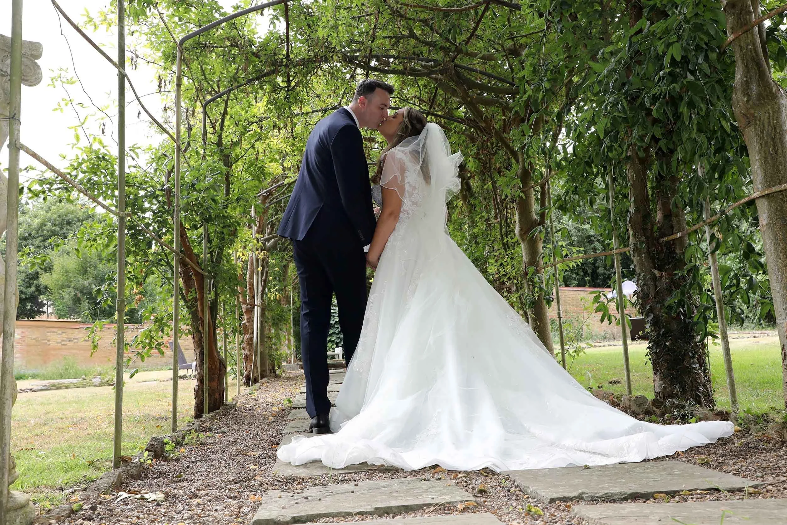 A bride and groom sharing a kiss outdoors under a green, leafy canopy during their wedding ceremony.