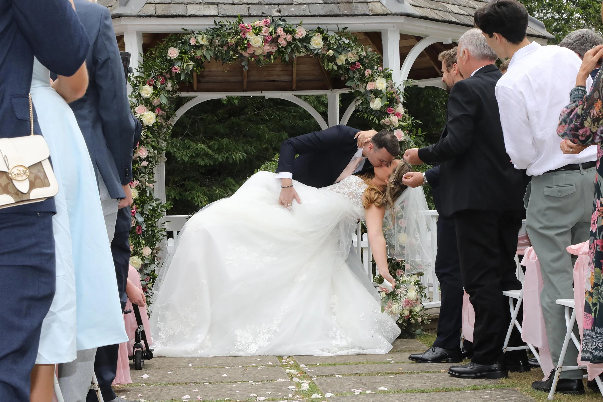 A bride and groom kiss during their outdoor wedding ceremony beneath an arch decorated with pink and white roses and greenery, surrounded by seated guests.