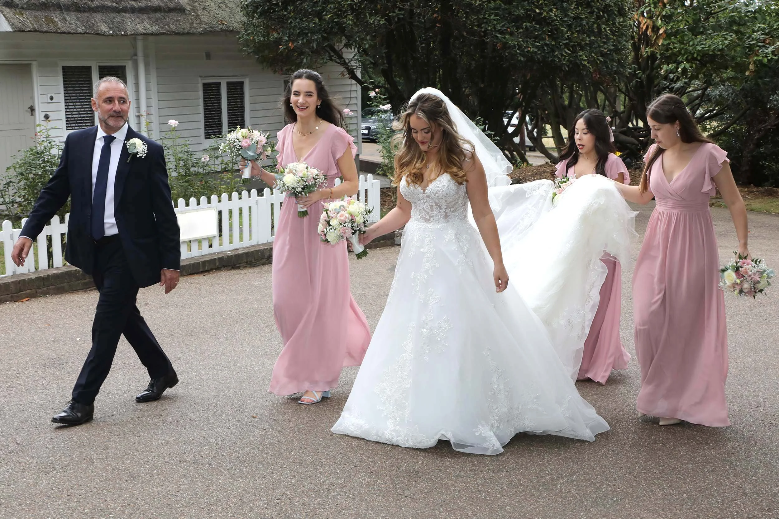 A bride in a white wedding dress walks alongside bridesmaids dressed in pink, holding bouquets, with an older man in a dark suit leading the group outdoors.