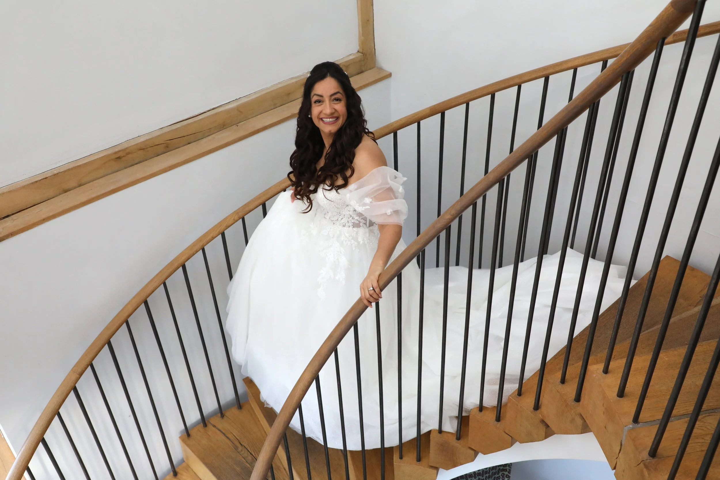A woman in a white wedding dress standing on a wooden staircase with black metal railings, smiling at the camera.