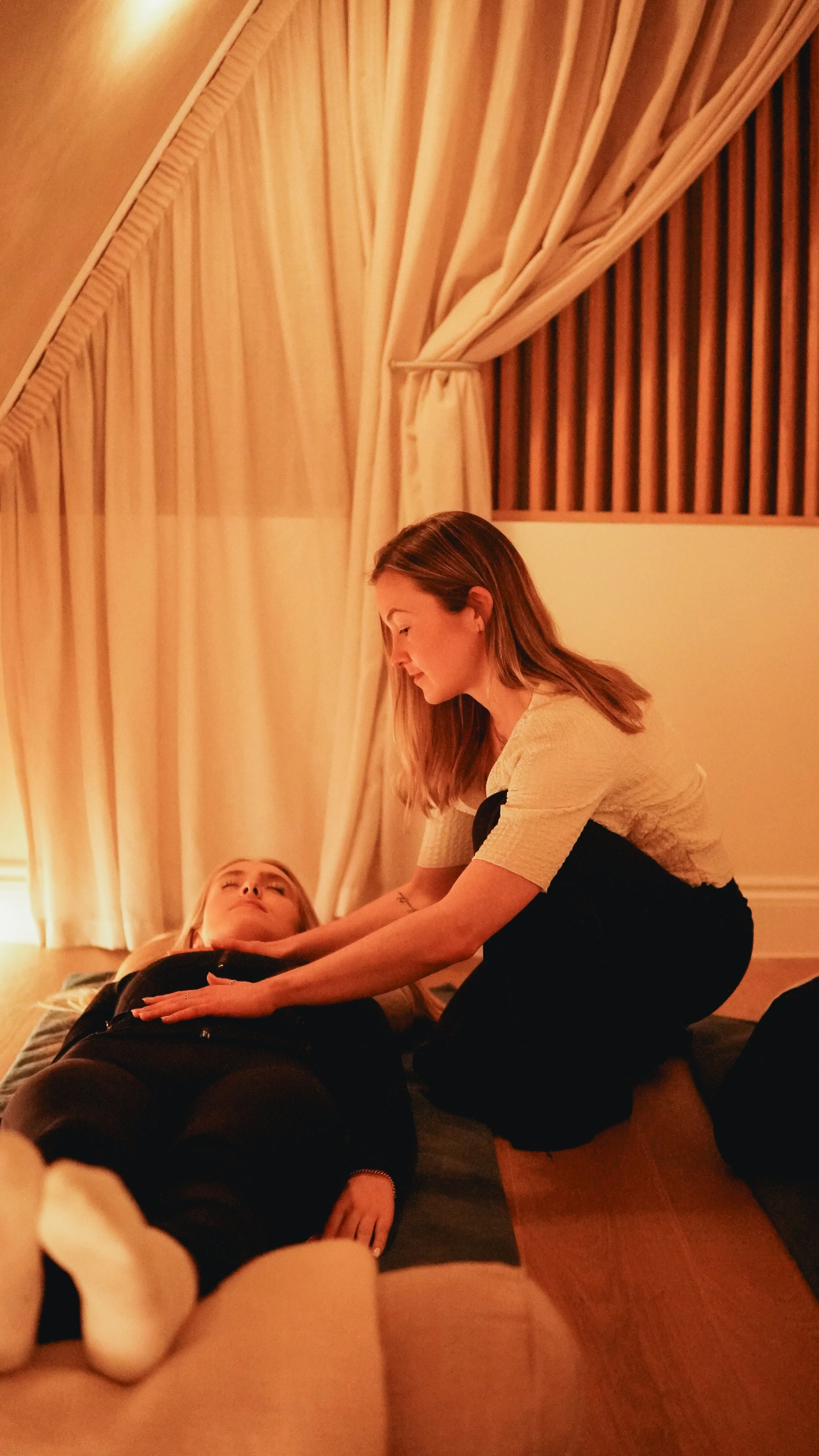 A woman providing a massage to a woman lying on a mat in a cozy, softly lit room with curtains.