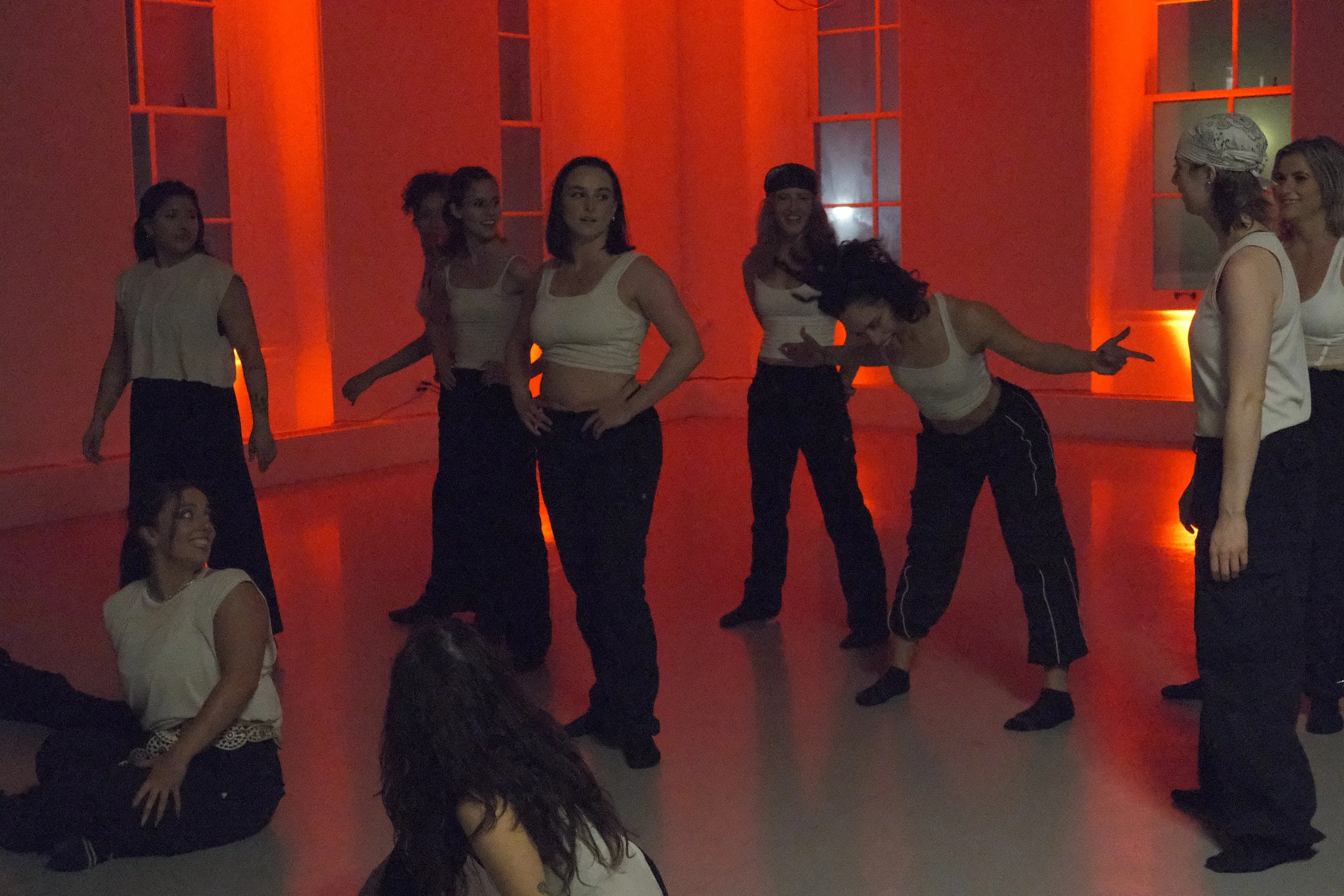 A woman practicing a high kick and balancing on one foot in a room with orange lighting, with a person filming her and a window in the background.