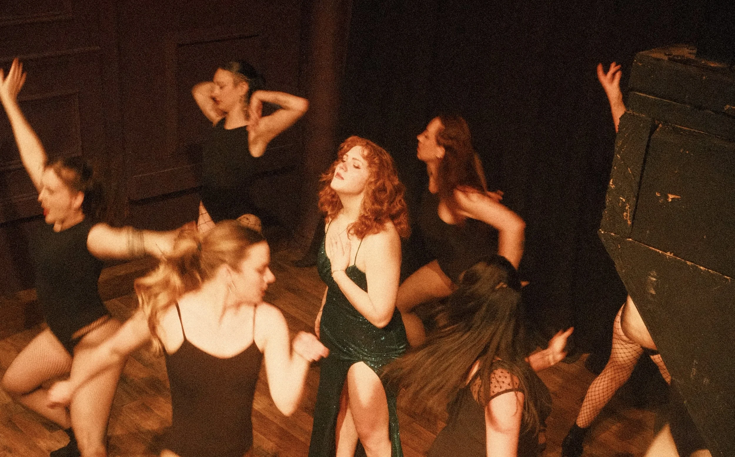 A woman with curly red hair wearing a green sparkly dress, surrounded by several women with outstretched hands, against a black curtain background.