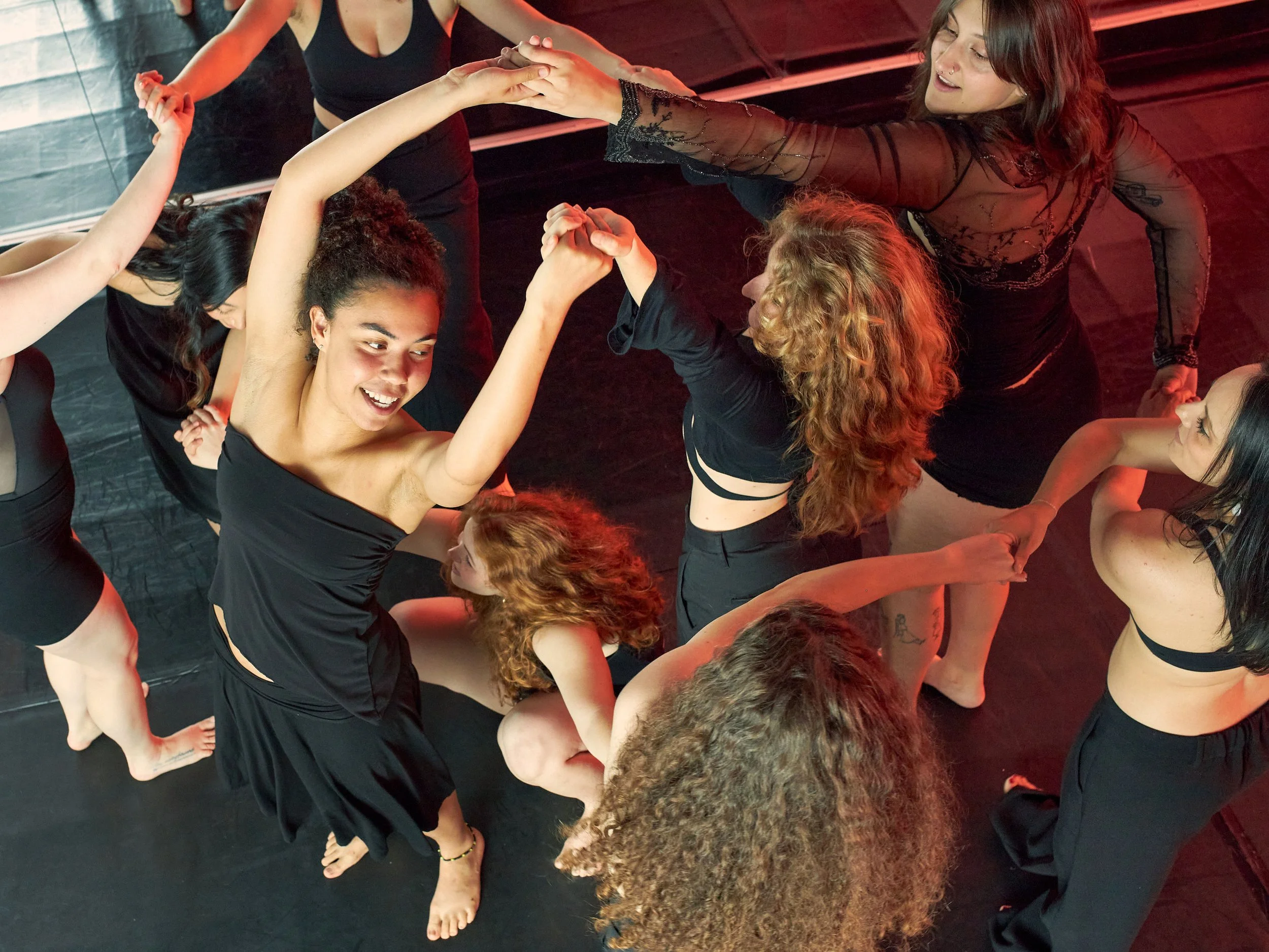 A group of women with different hair colors and styles are engaged in an activity, holding hands and forming a circle on a black floor in a dimly lit room with a red hue, possibly participating in a dance or yoga class.