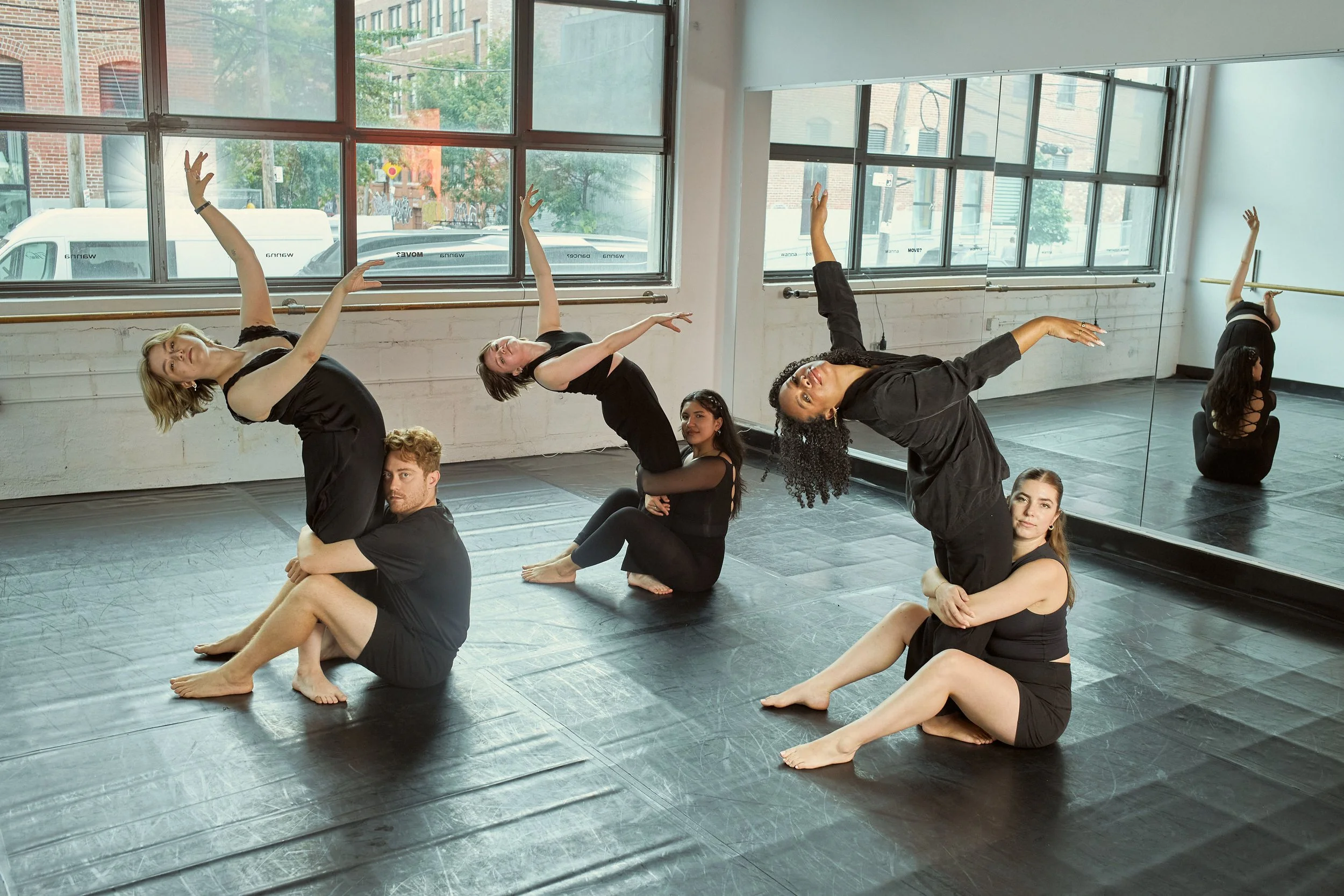 A group of dancers practicing in a studio with large windows and a mirror, some dancers seated on the floor and others in various dance poses.