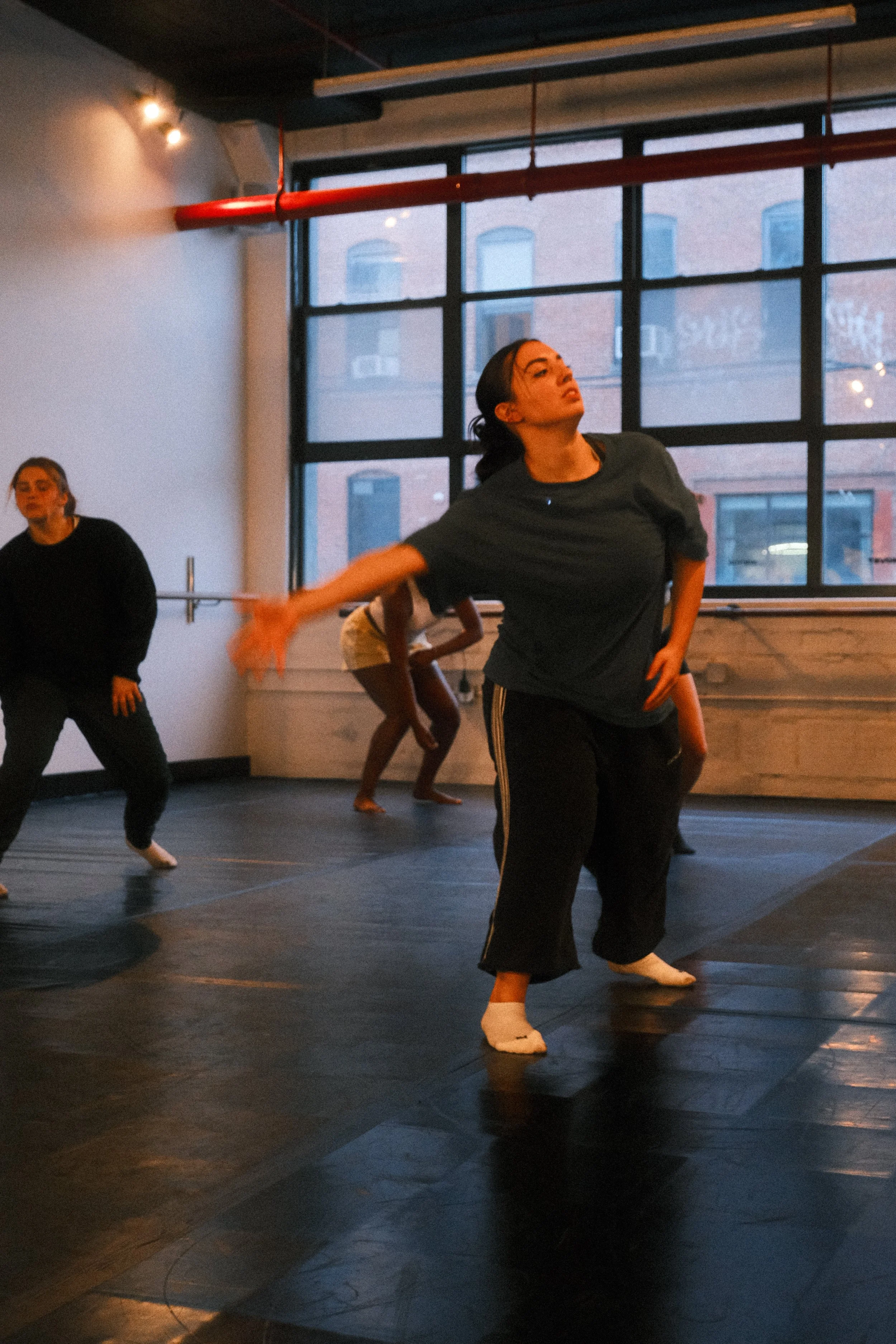 Two women dance in front of a white brick wall with red abstract art, one in black athletic wear and the other in loose gray top and black pants.