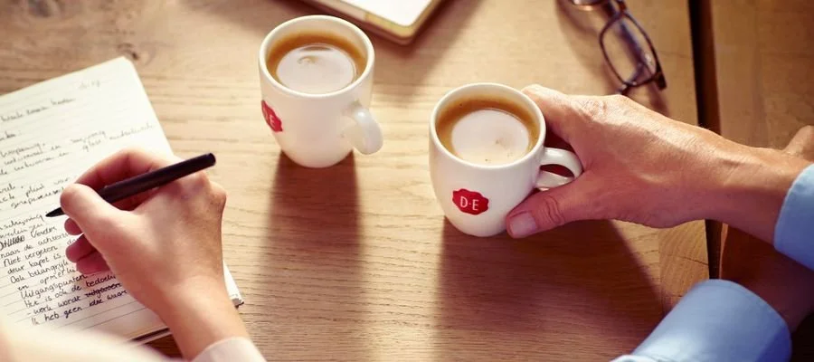 Two people sitting at a wooden table, one writing in a notebook and the other holding a small coffee cup with the logo 'DE', short for Douwe Egberts.