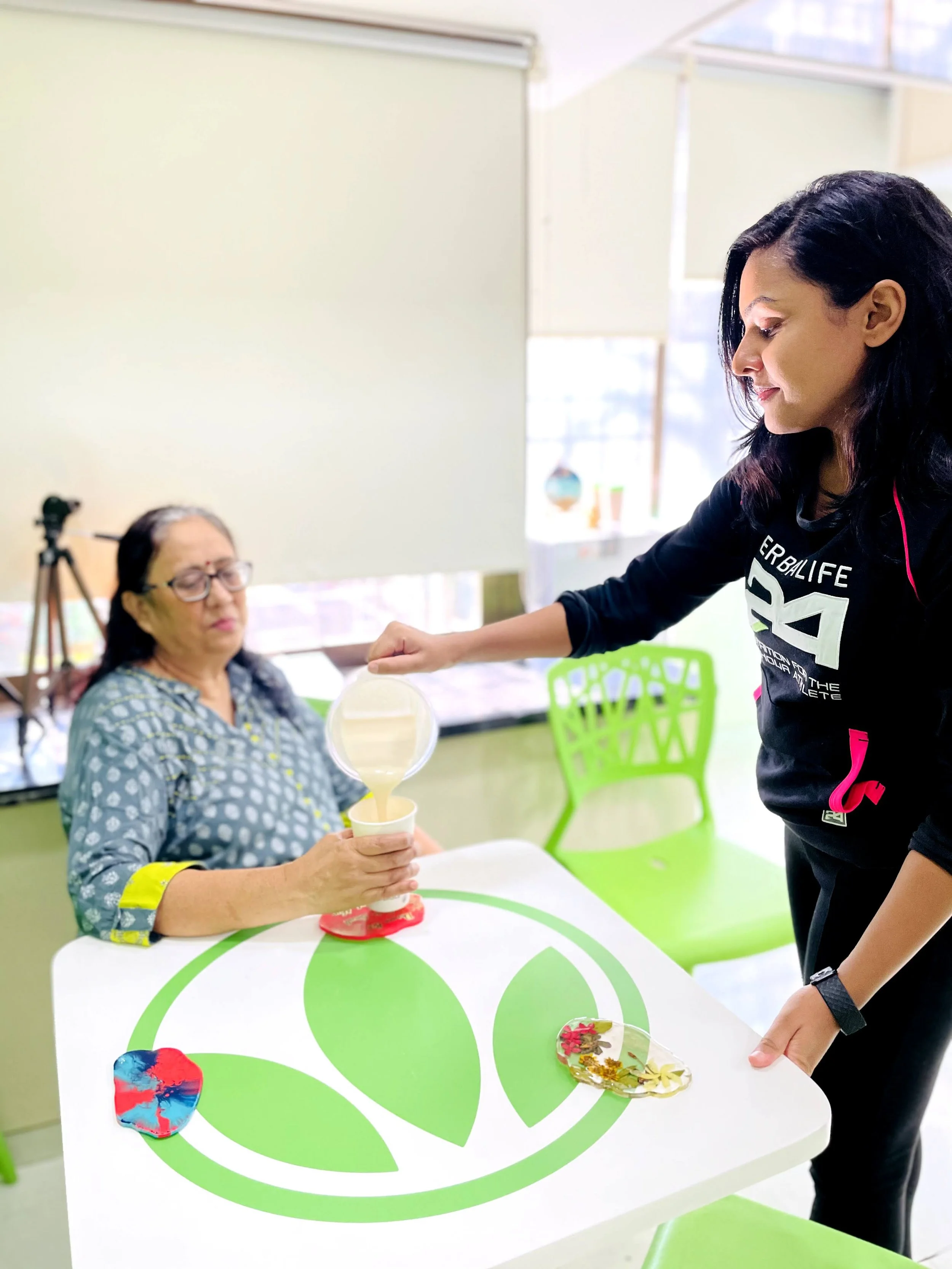A young woman in a black Herbalife shirt is pouring a creamy nutritional shake into a cup for an older woman with glasses, sitting at a white table with green leaf motifs. The room has bright natural light and green chairs.