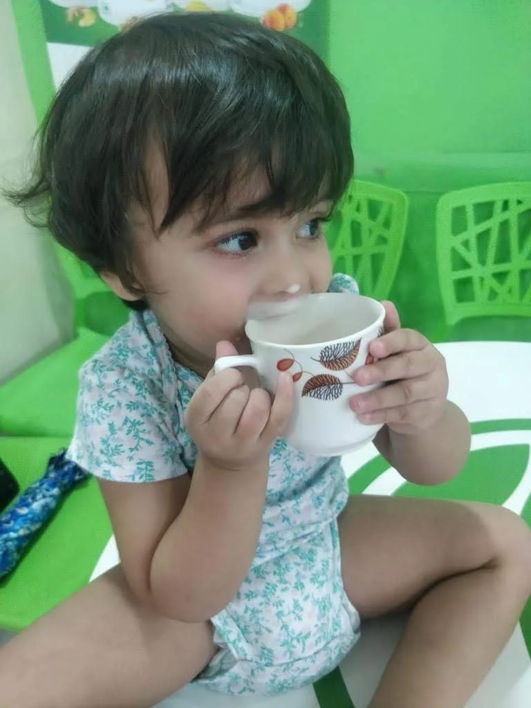 A young child with dark hair is sitting at a table, holding a mug with a leaf pattern, and appears to be drinking from it. The background includes green chairs and decor.
