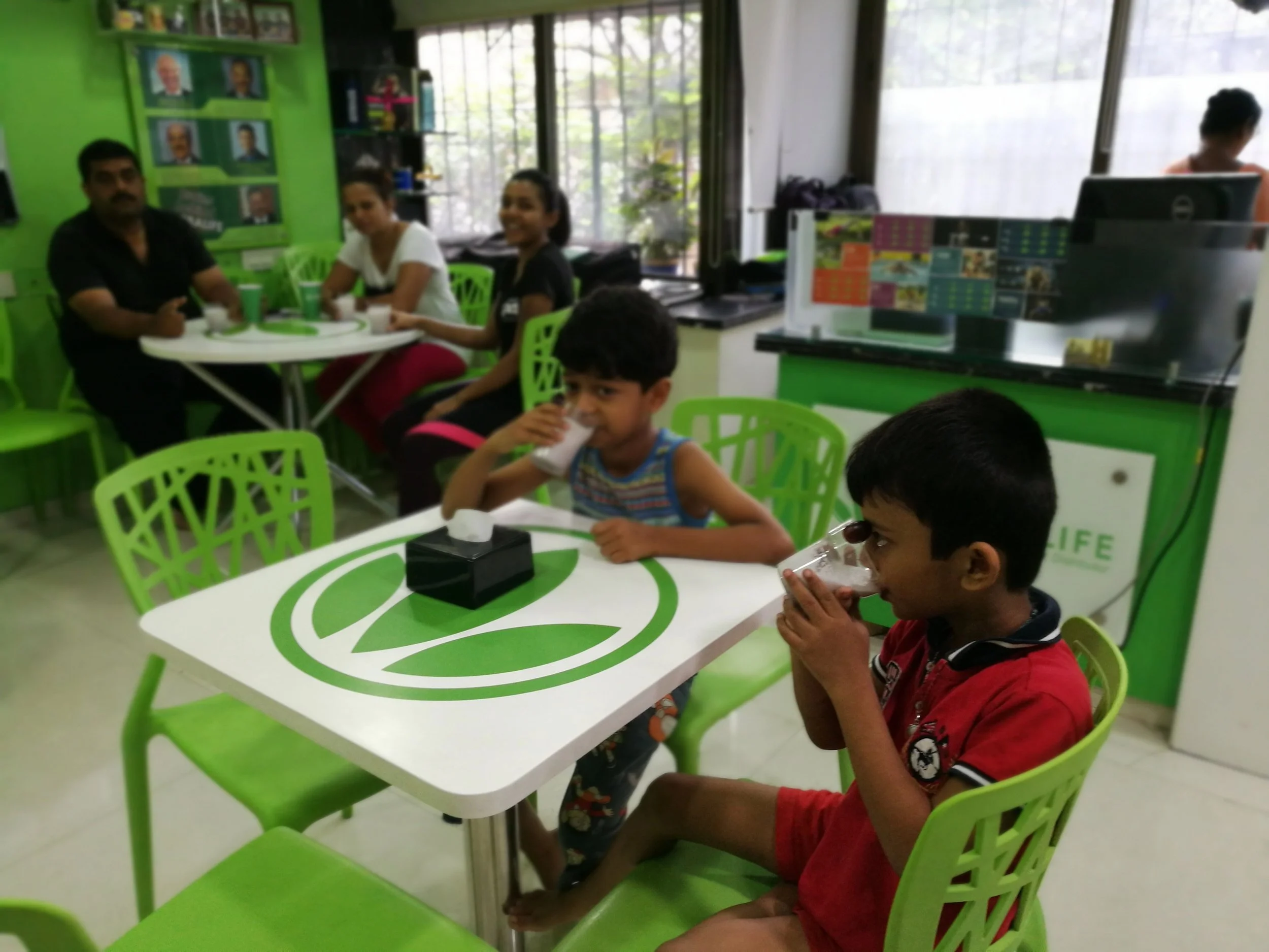 Two young boys sitting at a white table with a green logo, drinking from glasses, in a brightly lit cafe with green chairs and walls. In the background, a family of four is seated at a different table, and a woman is working near the counter.